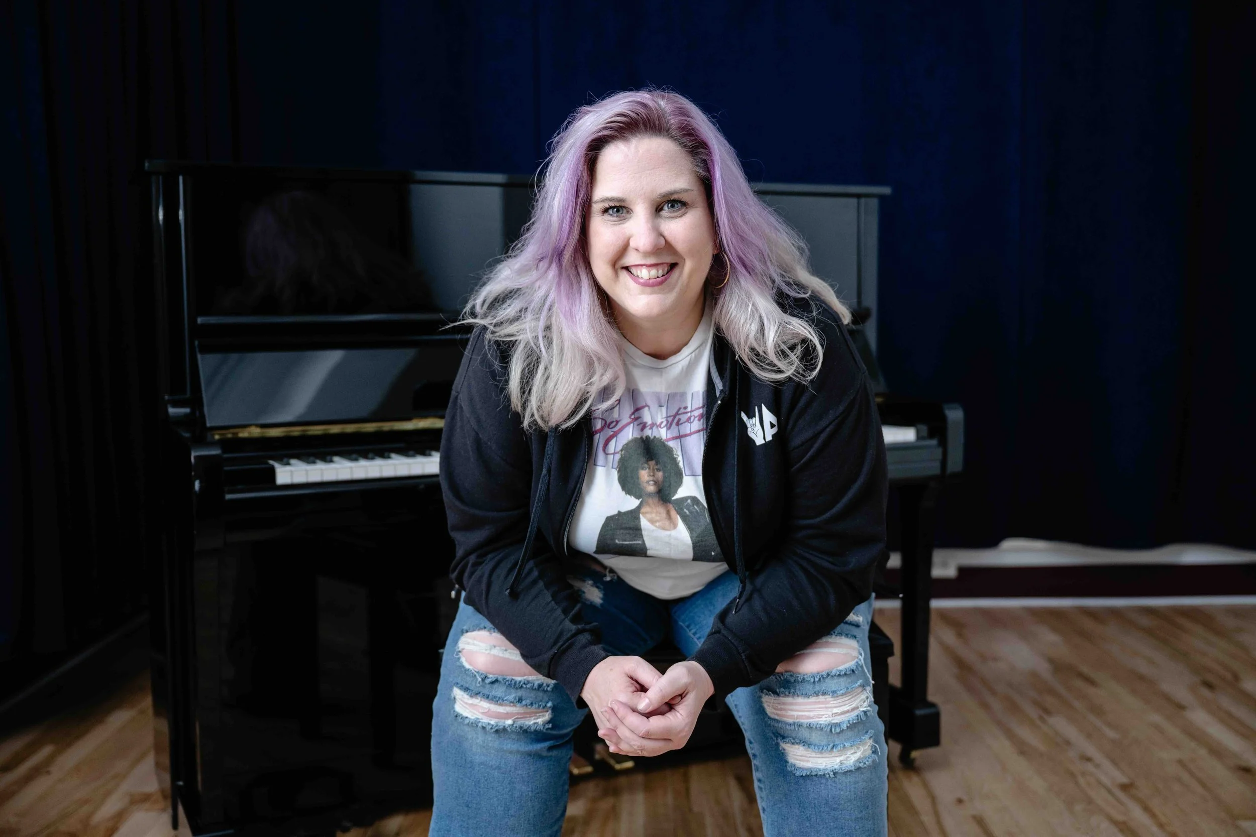 A woman with purple and blonde hair smiling at the camera, sitting in front of a black piano, wearing a graphic t-shirt and ripped jeans.