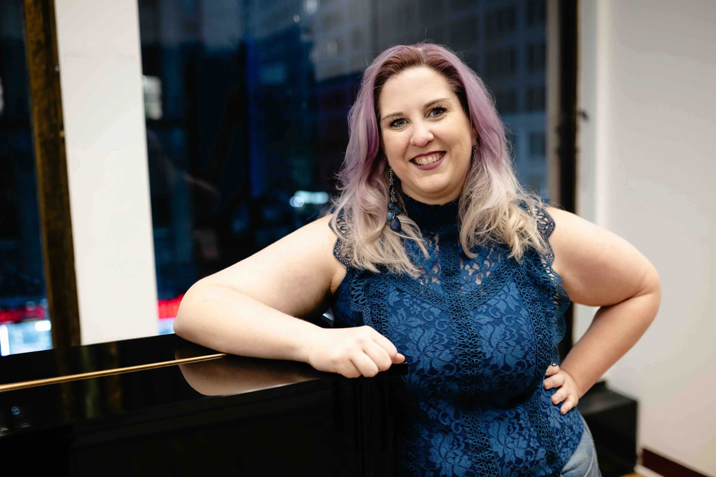 Shannah Rae is a woman with purple and blonde hair smiling, posing with her arm resting on a black grand piano inside a room with large windows at night.