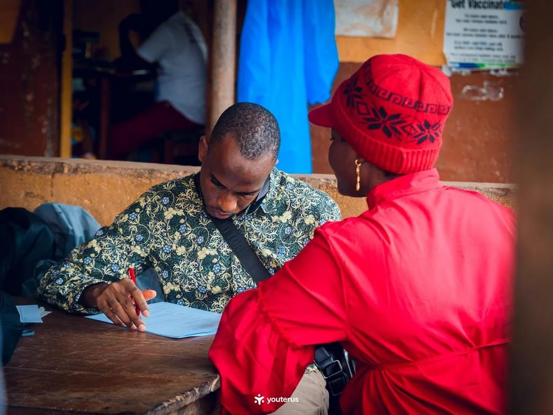 A healthcare worker and a woman perform a medical examination on a pregnant woman lying on a bed in an indoor setting, with a poster about sexual health on the wall behind them.