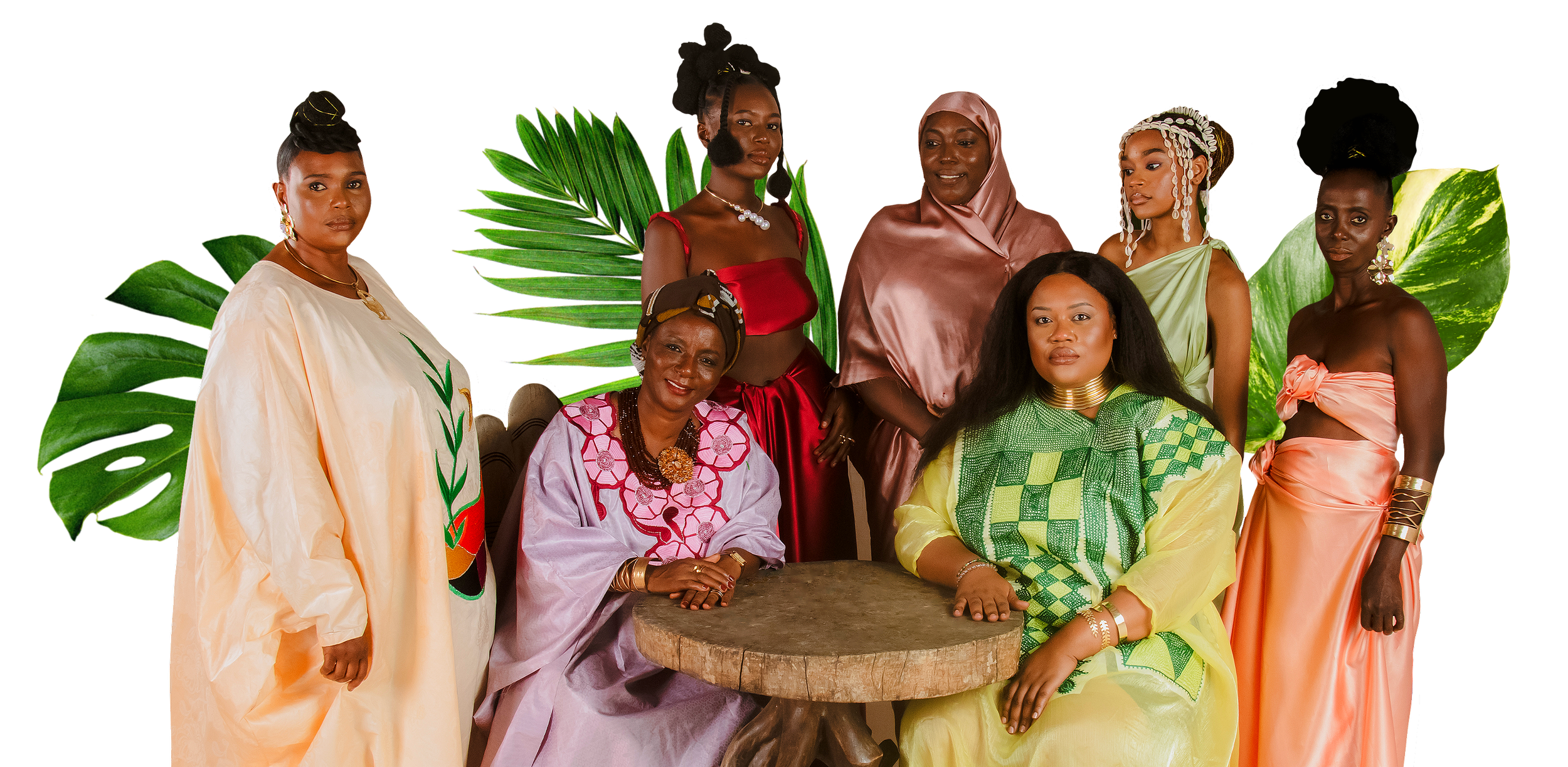 Group of seven women in colorful traditional and modern African attire posing around a wooden table with green tropical leaves in the background.