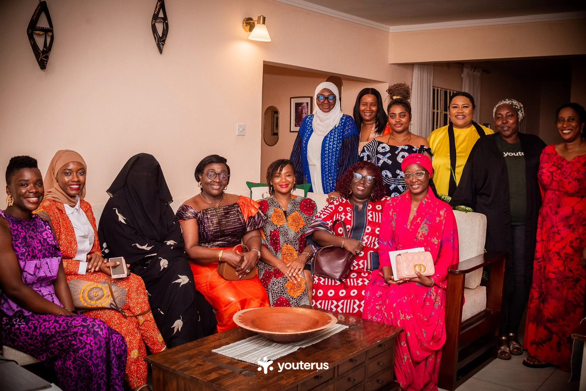 A group of fourteen women gathered together in a living room, smiling for a photo. They are dressed in colorful and patterned traditional and modern clothing, with a few holding bags or accessories. The background shows a beige wall with decorative wall pieces and framed photos.