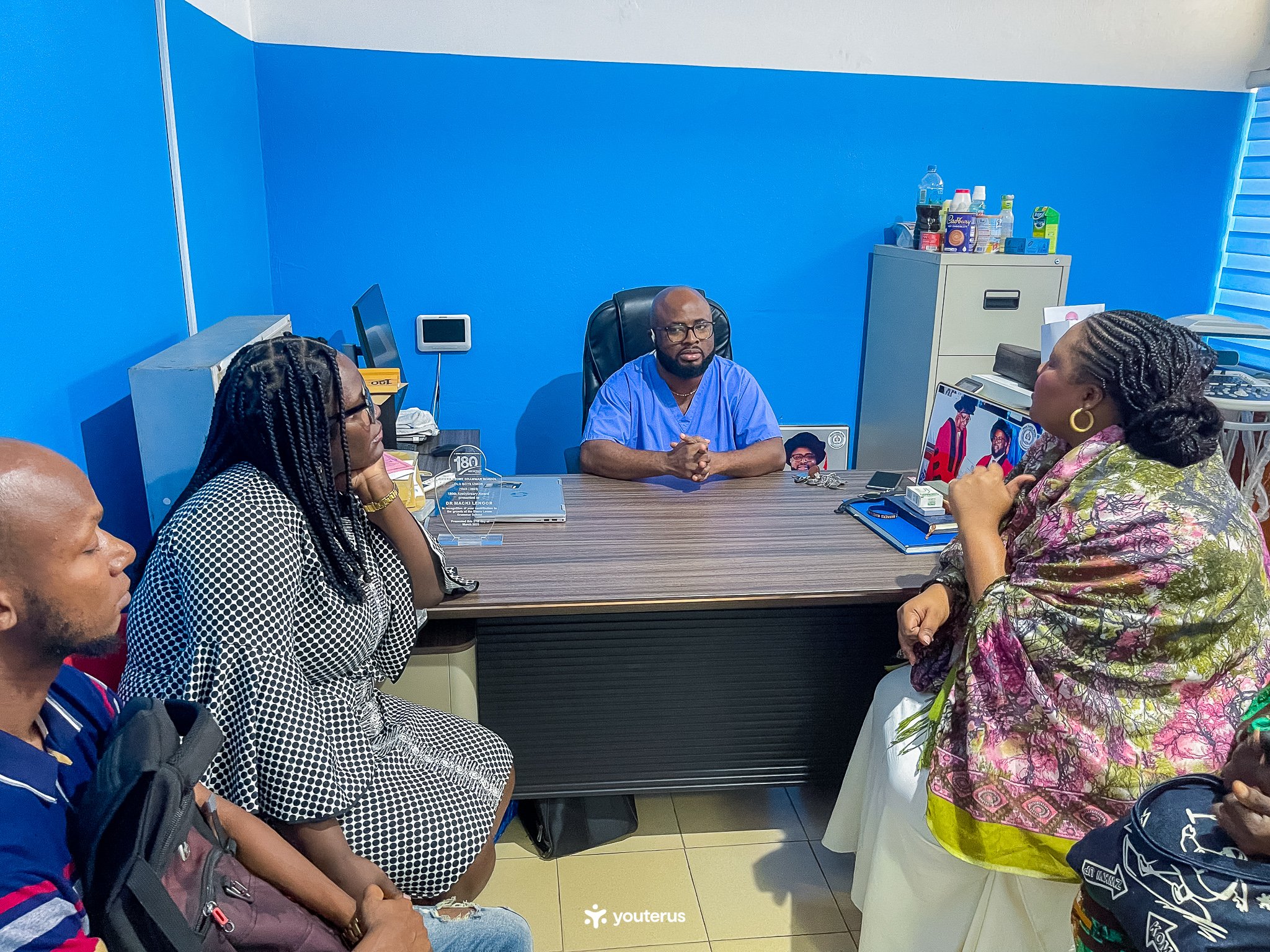A group of four people engaged in a discussion with a doctor in an office. The doctor sits behind a desk, wearing blue scrubs, and the others are seated in front of him. The office has a blue wall, a filing cabinet, and various office supplies.