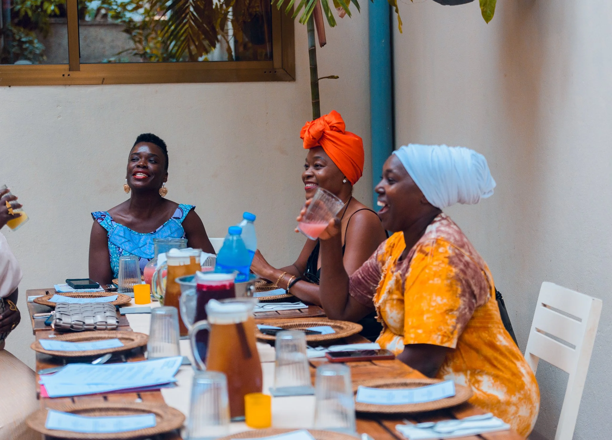 Three women enjoying a meal and drinks at a dining table during a social gathering, with one woman wearing a bright blue dress, another in a black top with an orange head wrap, and the third in a patterned dress with a white head wrap.