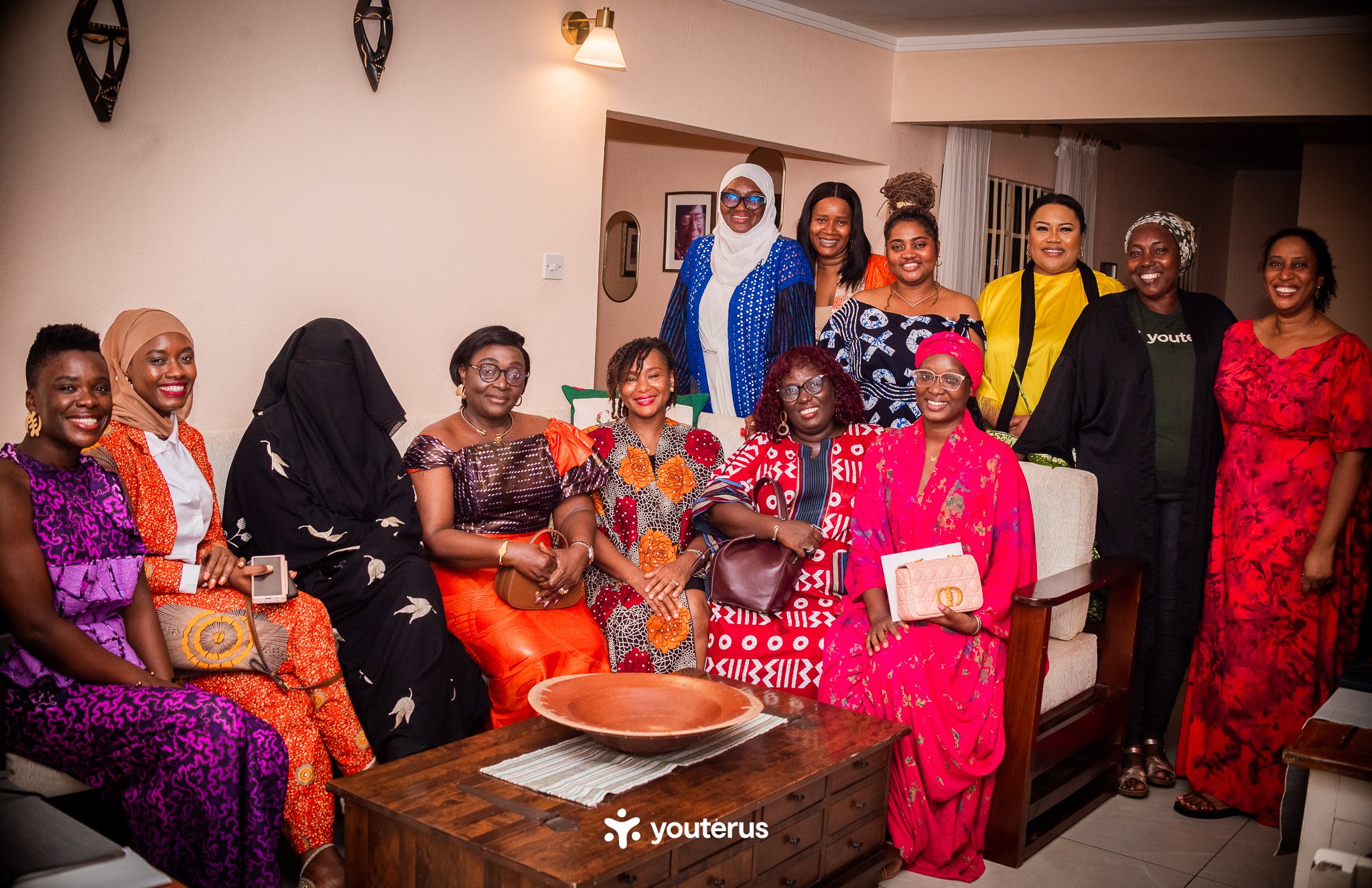 A group of women posing indoors for a photo, some sitting on a sofa and others standing behind it, dressed in colorful traditional and modern attire, smiling at the camera.