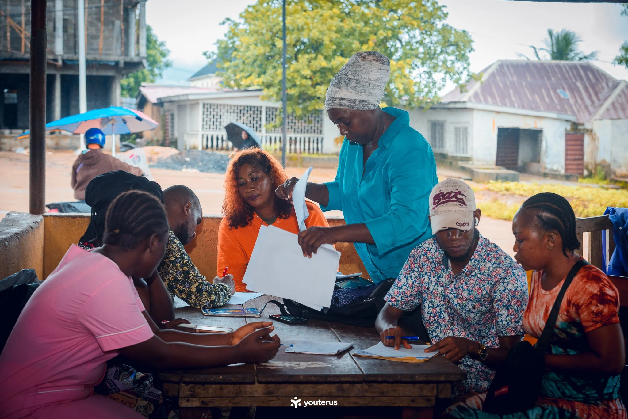 Group of people sitting around a table outdoors, with a woman in a blue shirt distributing papers and others taking notes.