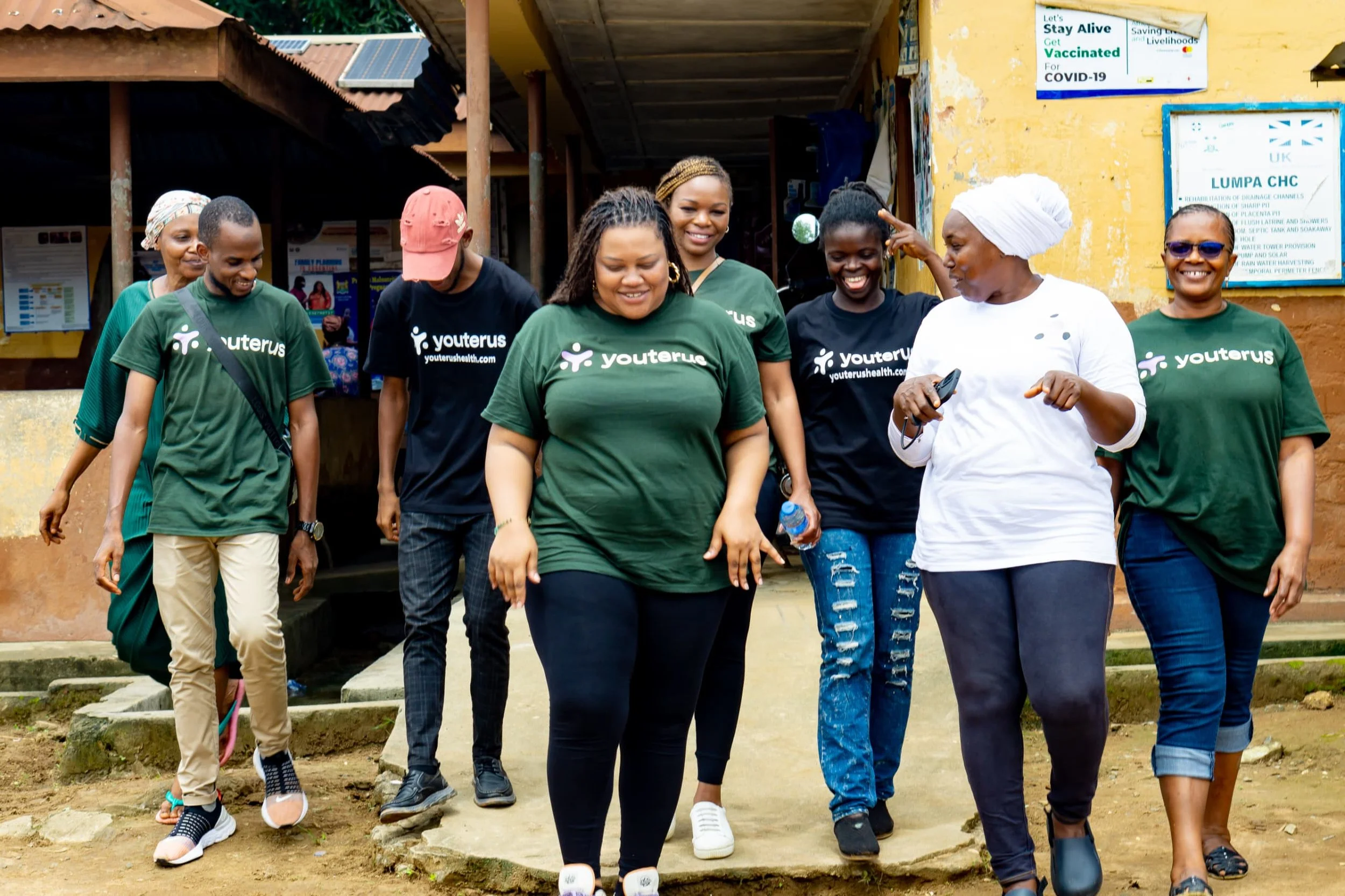 Group of women walking and smiling outside a building, some wearing 'Youterus' t-shirts, with one woman in a white headscarf and white t-shirt gesturing and holding a phone, in a casual outdoor setting.