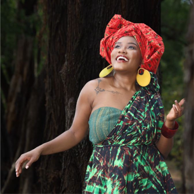 A woman with a bright smile wearing traditional African clothing and jewelry, posing outdoors against a tree.