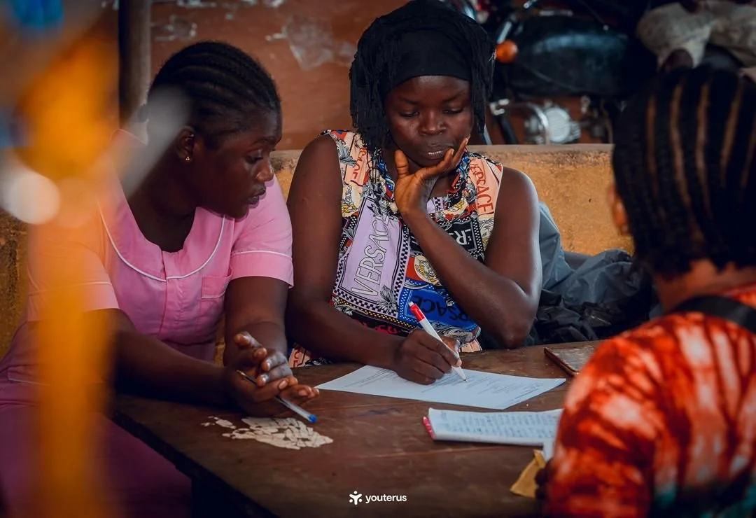 Three women sit around a wooden table, focused on writing and discussing documents.