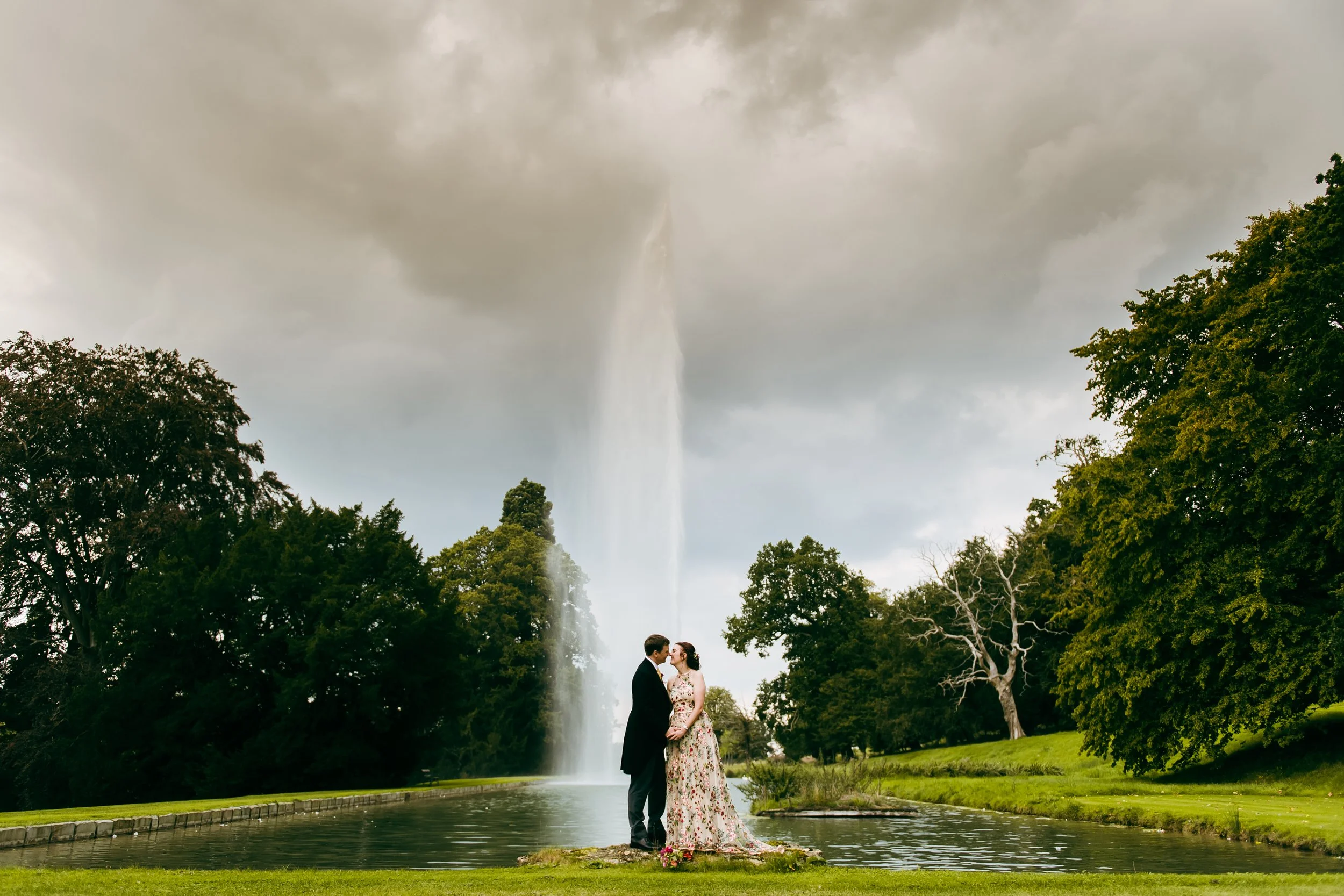 A couple in wedding attire standing on a small patch of grass in front of a pond, with a tall fountain behind them in the Cotswolds, surrounded by green trees and gray cloudy sky.