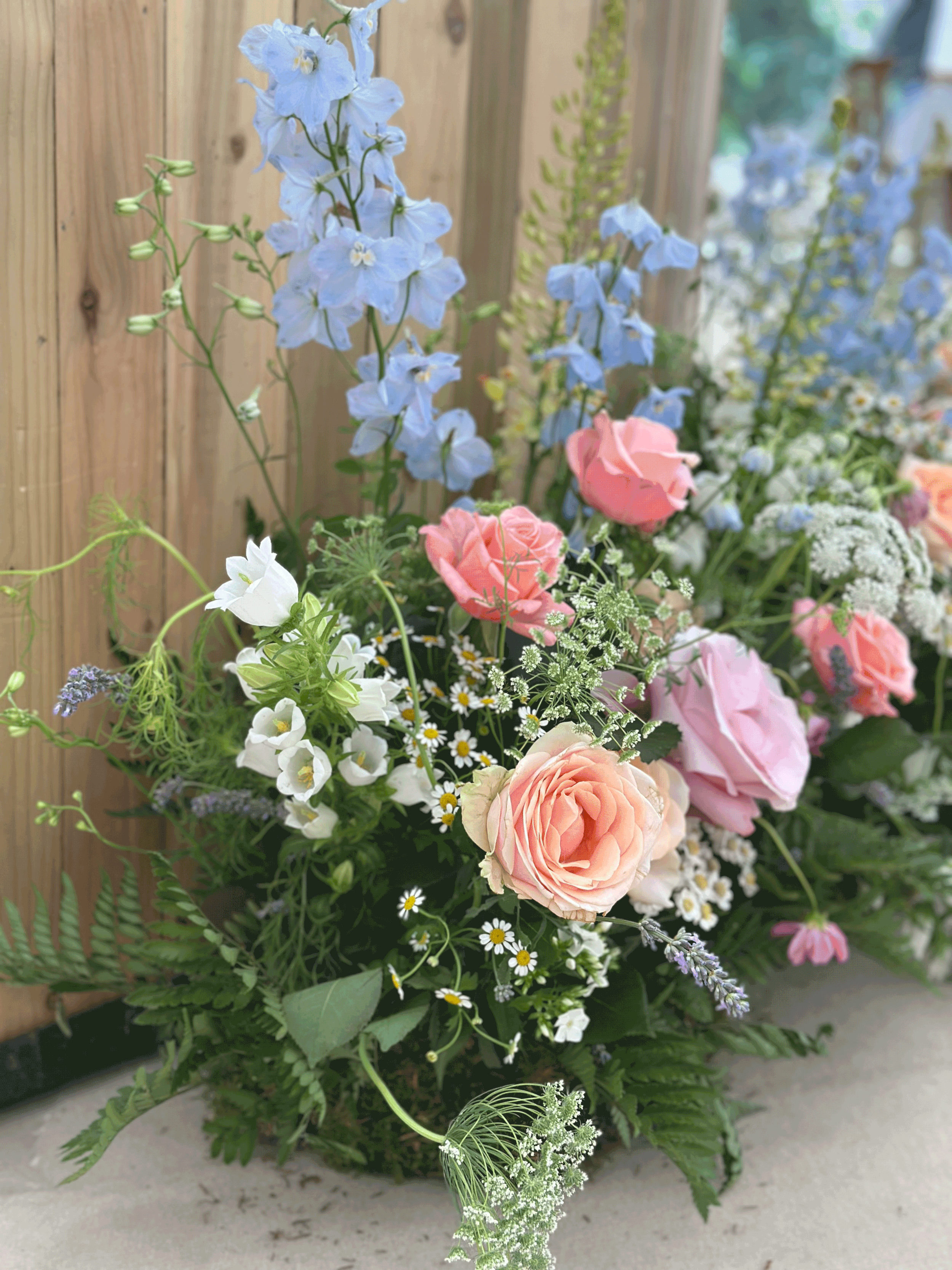 A colorful flower arrangement with pink roses, blue delphiniums, white daisies, and other small white and purple flowers, set against a wooden panel background in the Cotswolds