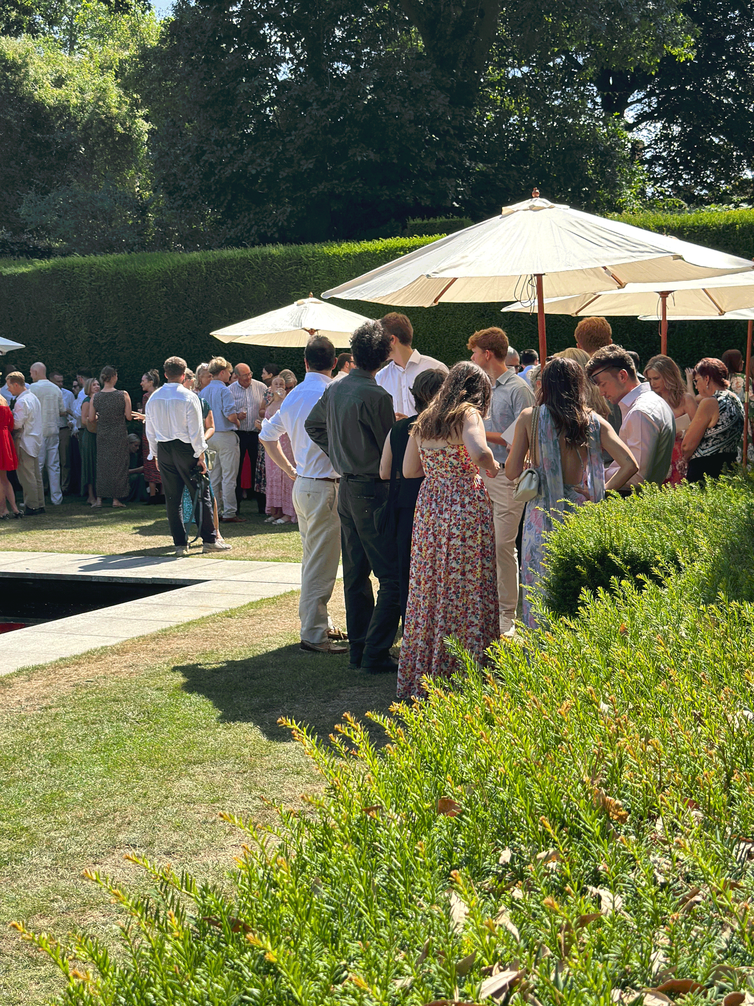 People gathered outdoors under umbrellas at a garden party or social event on a sunny day in the cotswolds
