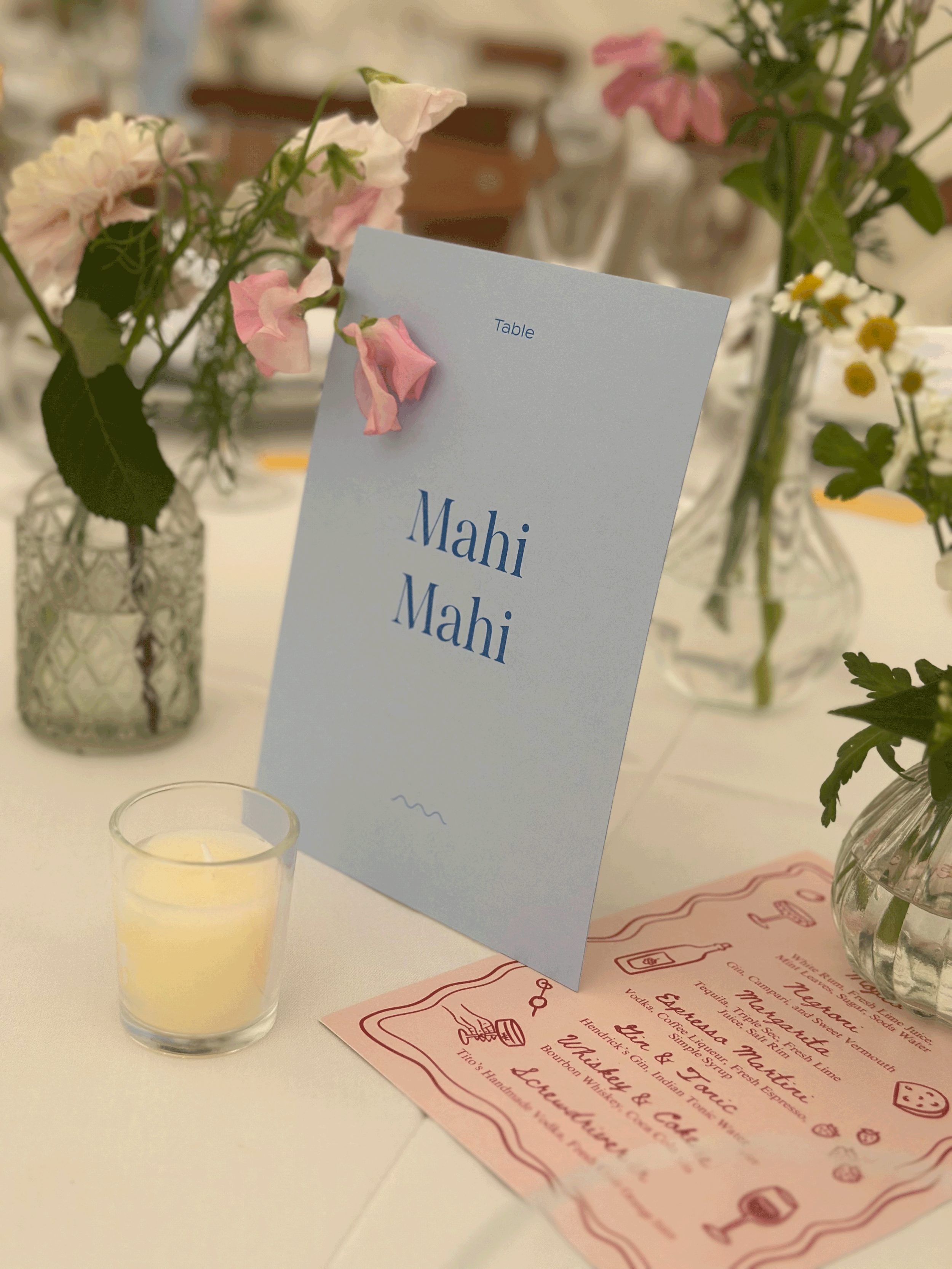 A wedding or event table setting with a light blue menu card labeled 'Mahi Mahi' in blue text, surrounded by vases of pink and white flowers, a small candle, and a pink and red menu on the table. In the Cotswolds