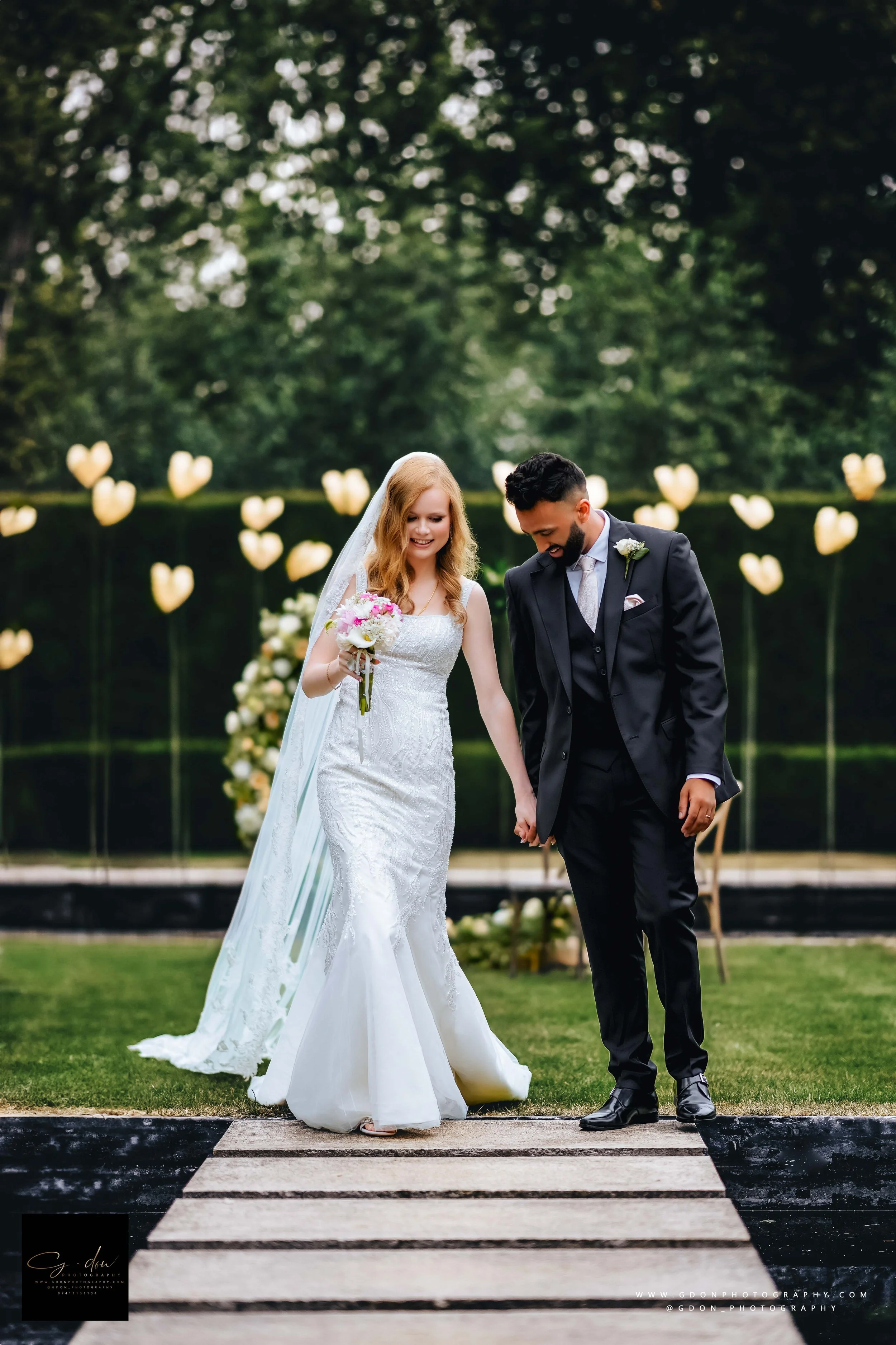 A bride and groom walking hand in hand on a wooden pathway outdoors, with heart-shaped string lights in the background, during their wedding celebration in the Cotswolds