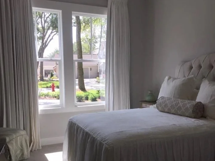 Bright bedroom with large window, white curtains, beige bed with pillows, and a nightstand with a small decorative object.