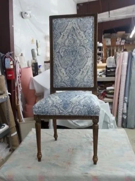 A vintage wooden chair with custom upholstered seat and backrest, featuring a blue and white patterned fabric, placed on a table in a workshop or storage area.