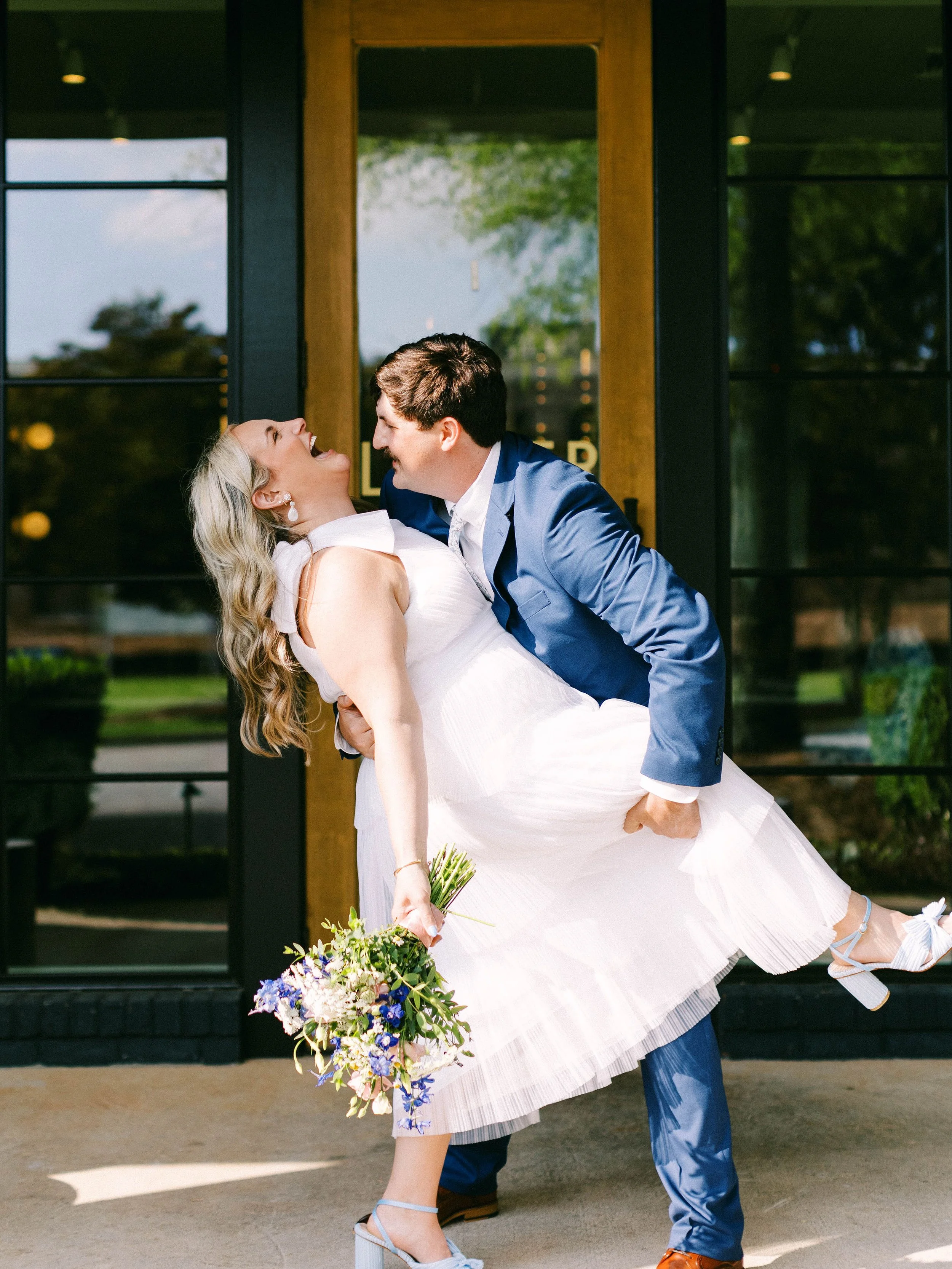 A groom in a blue suit holding a bride in a white dress outside a building, both laughing and enjoying a moment together.