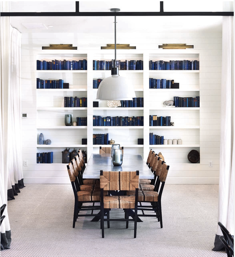 A dining room with a large wooden table surrounded by ten chairs with woven seats. Behind the table is a white wall with built-in shelves filled with blue books and decorative objects. A large pendant light hangs above the table.