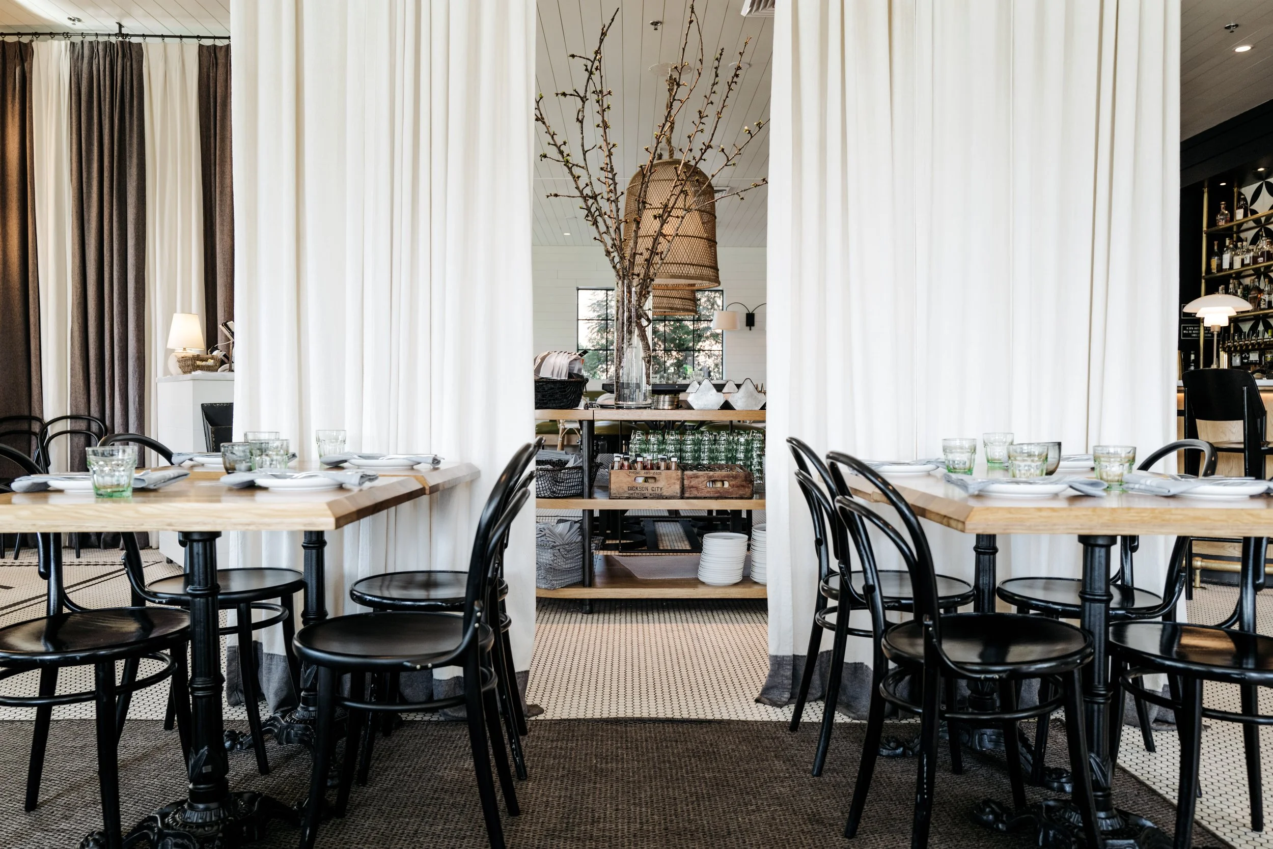 Interior of a restaurant with two wooden tables and black chairs, divided by white curtains, with a serving area in the background holding glassware, plates, and decorative branches in a large vase.