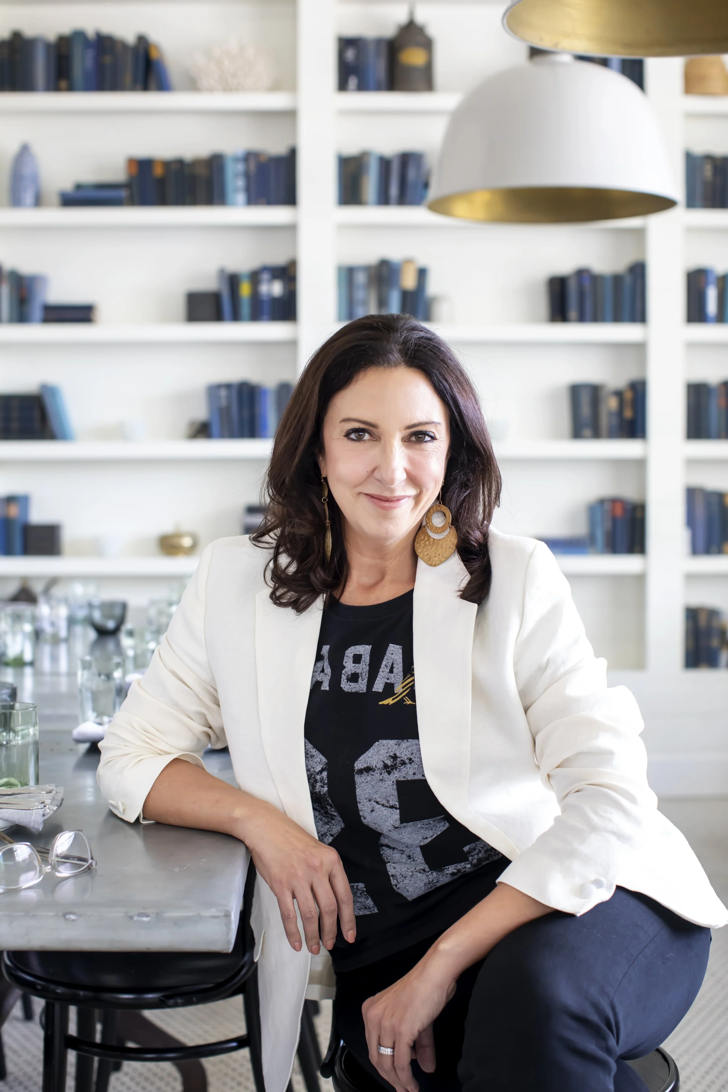 A woman with dark hair, wearing a black T-shirt with a graphic and a white blazer, sitting at a table in front of a bookshelf filled with books, smiling at the camera.