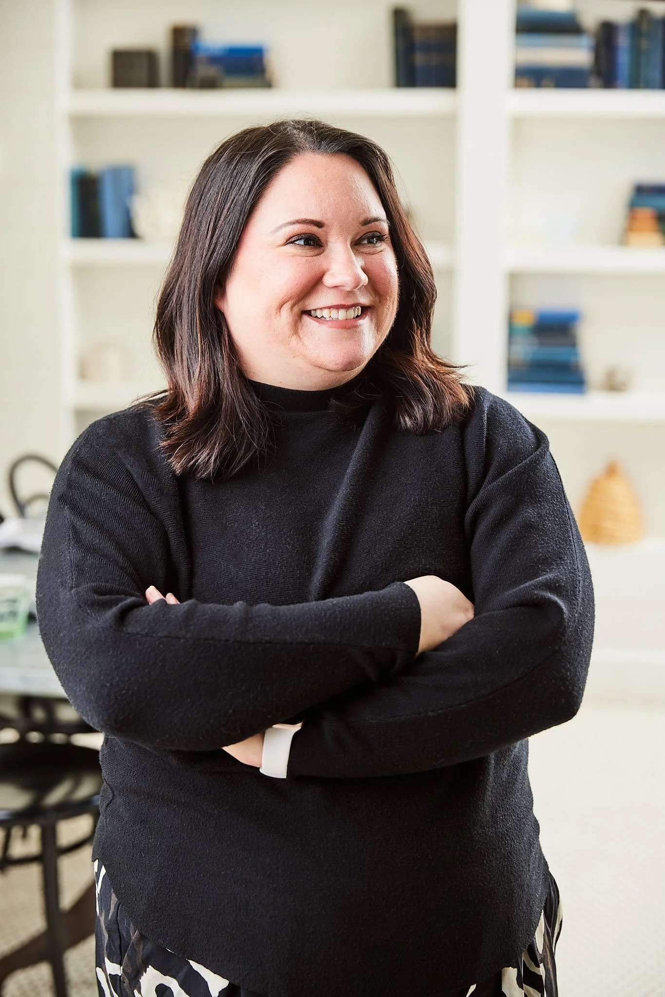 A woman with shoulder-length dark hair smiling and crossing her arms, standing in a room with bookshelves in the background.