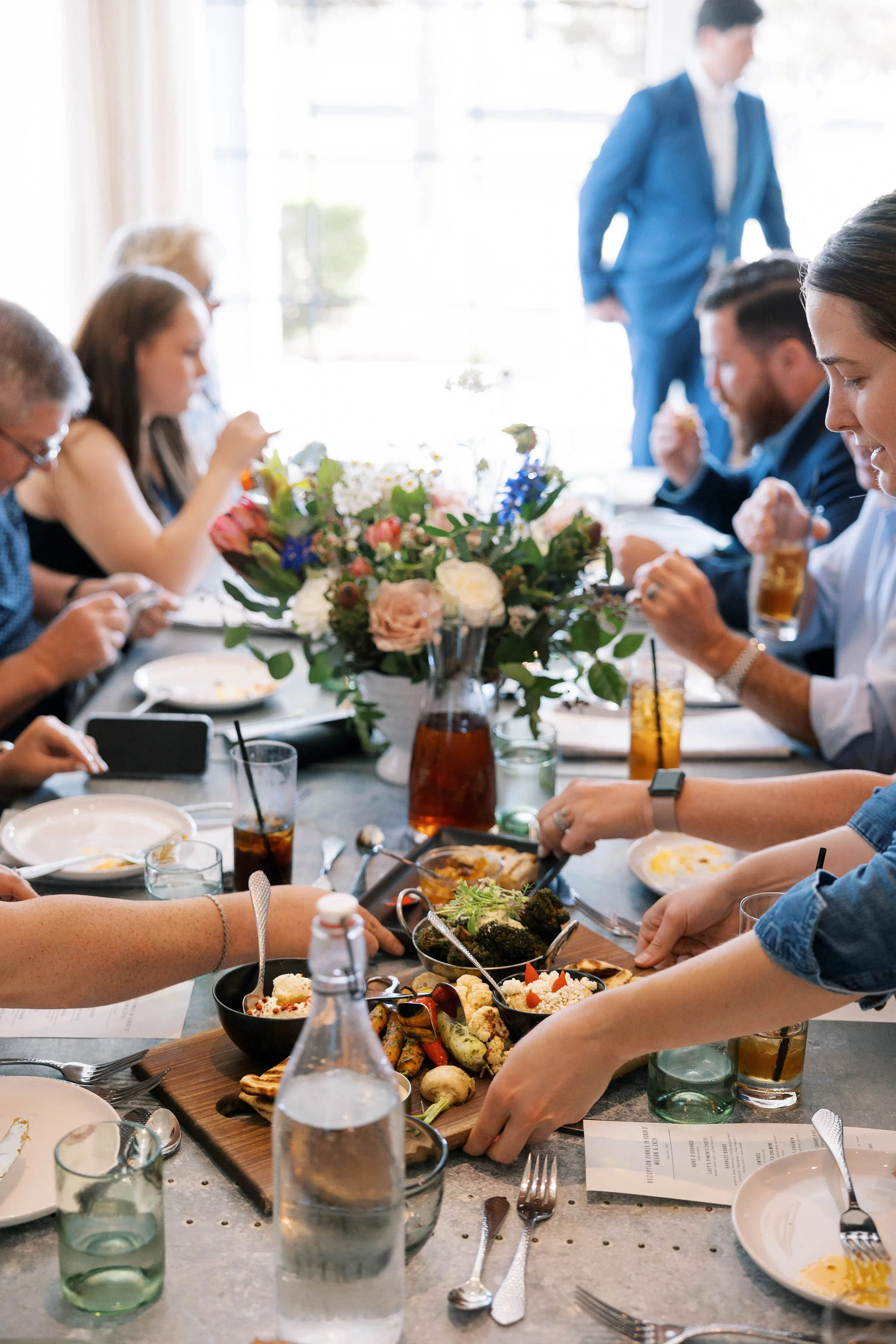 People gathered around a dining table enjoying a meal with a centerpiece of flowers, glasses of drinks, and assorted dishes.
