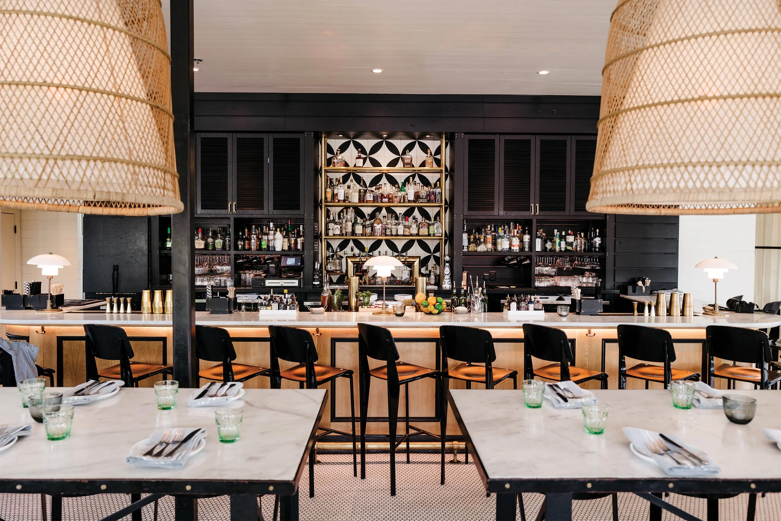 A stylish restaurant or bar interior with a marble table set with utensils and glasses in the foreground, black chairs around the table, and a bar area with bottles of alcohol and glassware behind it in the background, illuminated by warm lighting.