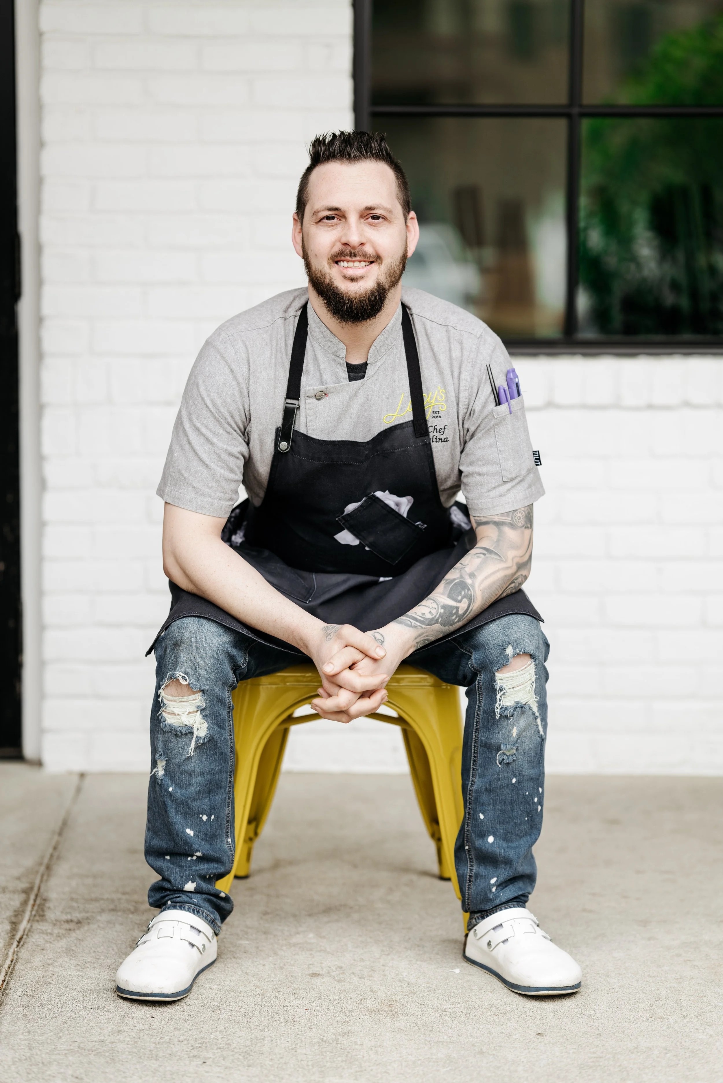 Male chef sitting on a yellow chair outside, wearing a gray chef coat, black apron, ripped jeans, and white shoes, with a tattooed arm and pens in his sleeve pocket.