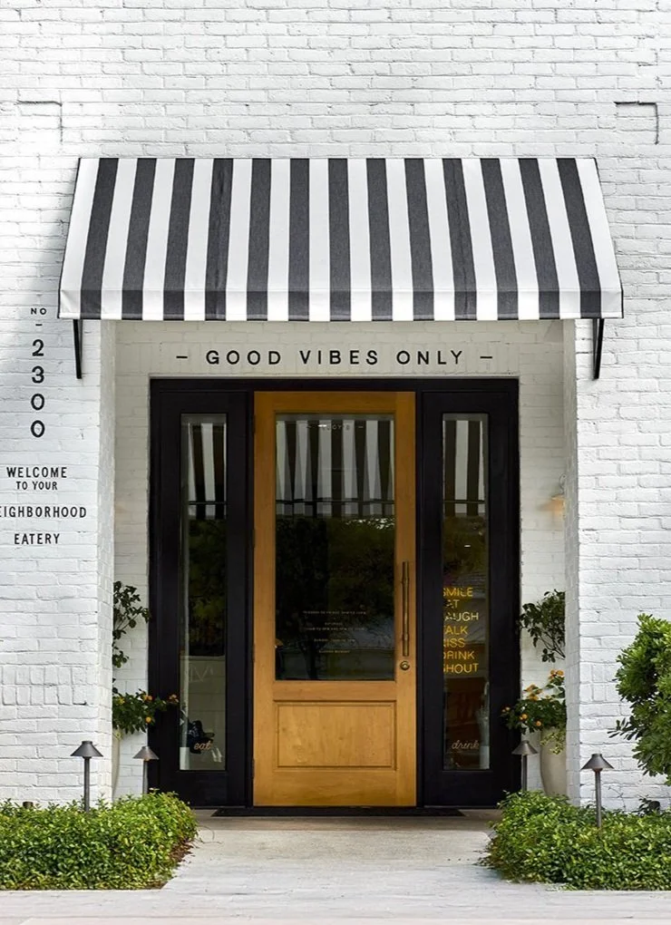 Exterior of a storefront with a black and white striped awning, white brick walls, and a wooden door. Above the door, there's a sign that reads 'GOOD VIBES ONLY.' To the side, there's a height measurement chart and a sign welcoming to the neighborhood eatery. There are small landscaped bushes and path lighting near the entrance.