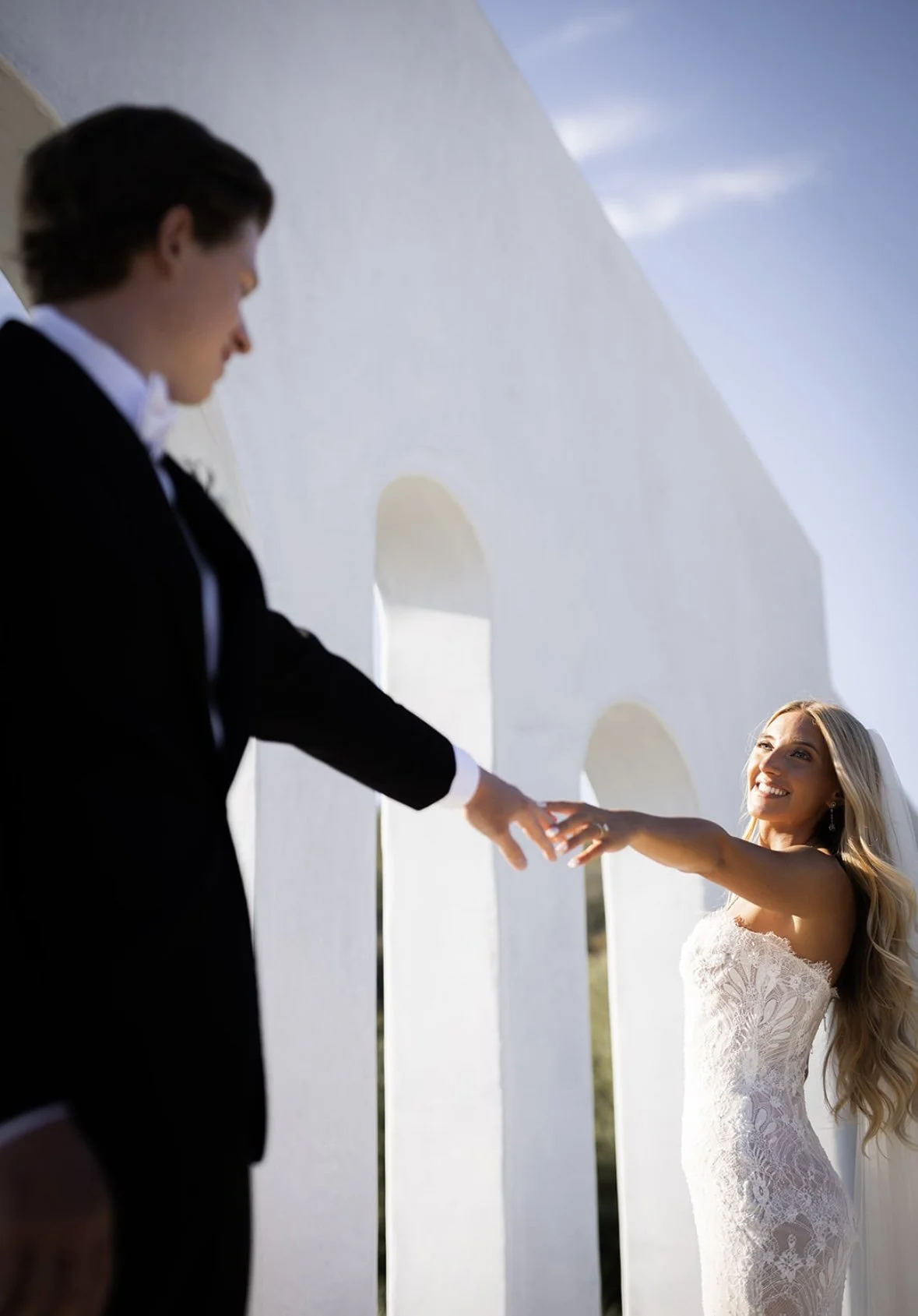 A bride and groom holding hands, standing outdoors near a white structure with arched openings, during a sunny day.