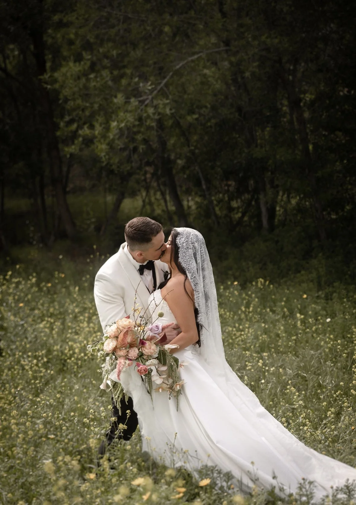 A bride and groom sharing a kiss in a field of small yellow flowers, with trees in the background. The bride is in a white gown holding a large bouquet, and the groom is in a white tuxedo with a black bow tie.