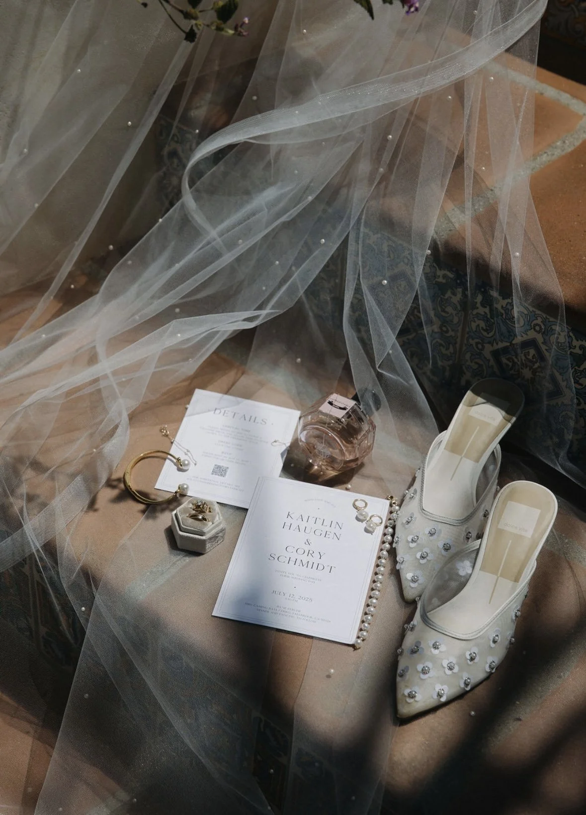 Wedding planning items including white embellished shoes, printed programs with names Kaitlin Haugen and Cory Schmidt, jewelry, perfume bottle, and a candle, arranged on a table with a sheer fabric overlay.