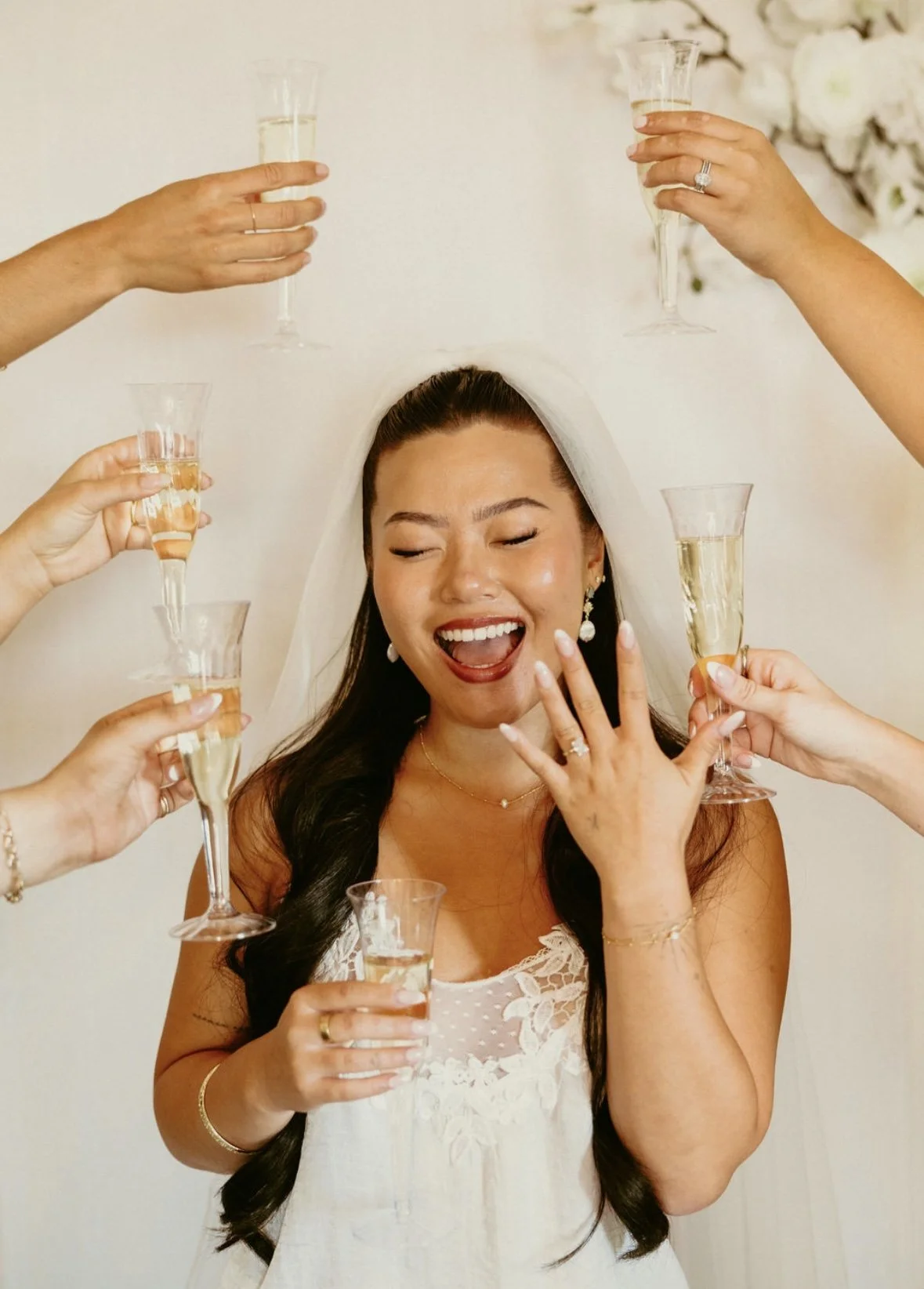 Bride celebrating with friends, raising glasses of champagne, smiling, with her hand showing a wedding ring.