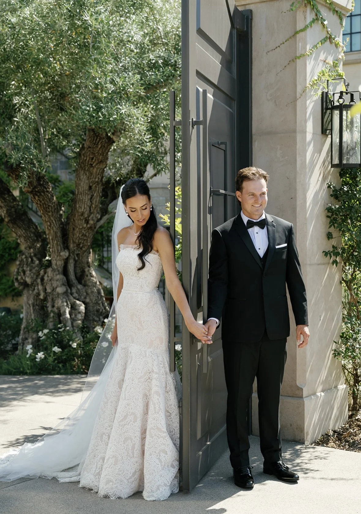 A bride in a white lace wedding dress holding hands with a groom in a black tuxedo, standing outside near an open gate with lush greenery in the background.