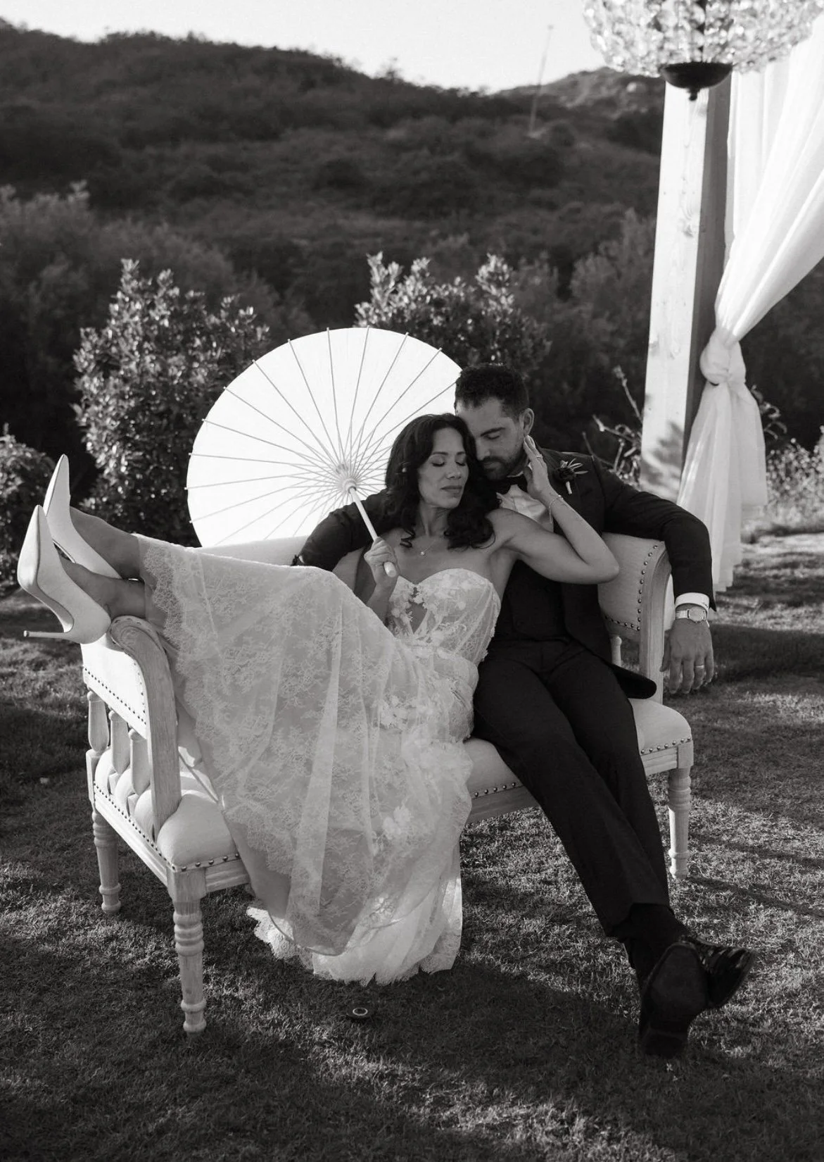 Black and white photo of a bride and groom sitting on a vintage sofa outdoors. The bride is holding a parasol and is dressed in a lace wedding gown, with her legs crossed on the sofa. The groom is dressed in a tuxedo. They appear relaxed and intimate, with a scenic landscape of trees and hills in the background.