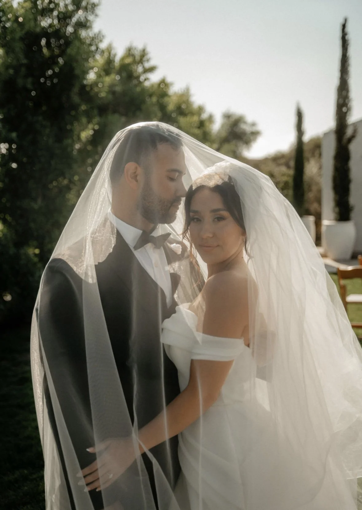 A bride and groom stand close together outdoors, under a sheer veil, with greenery and tall cypress trees in the background.