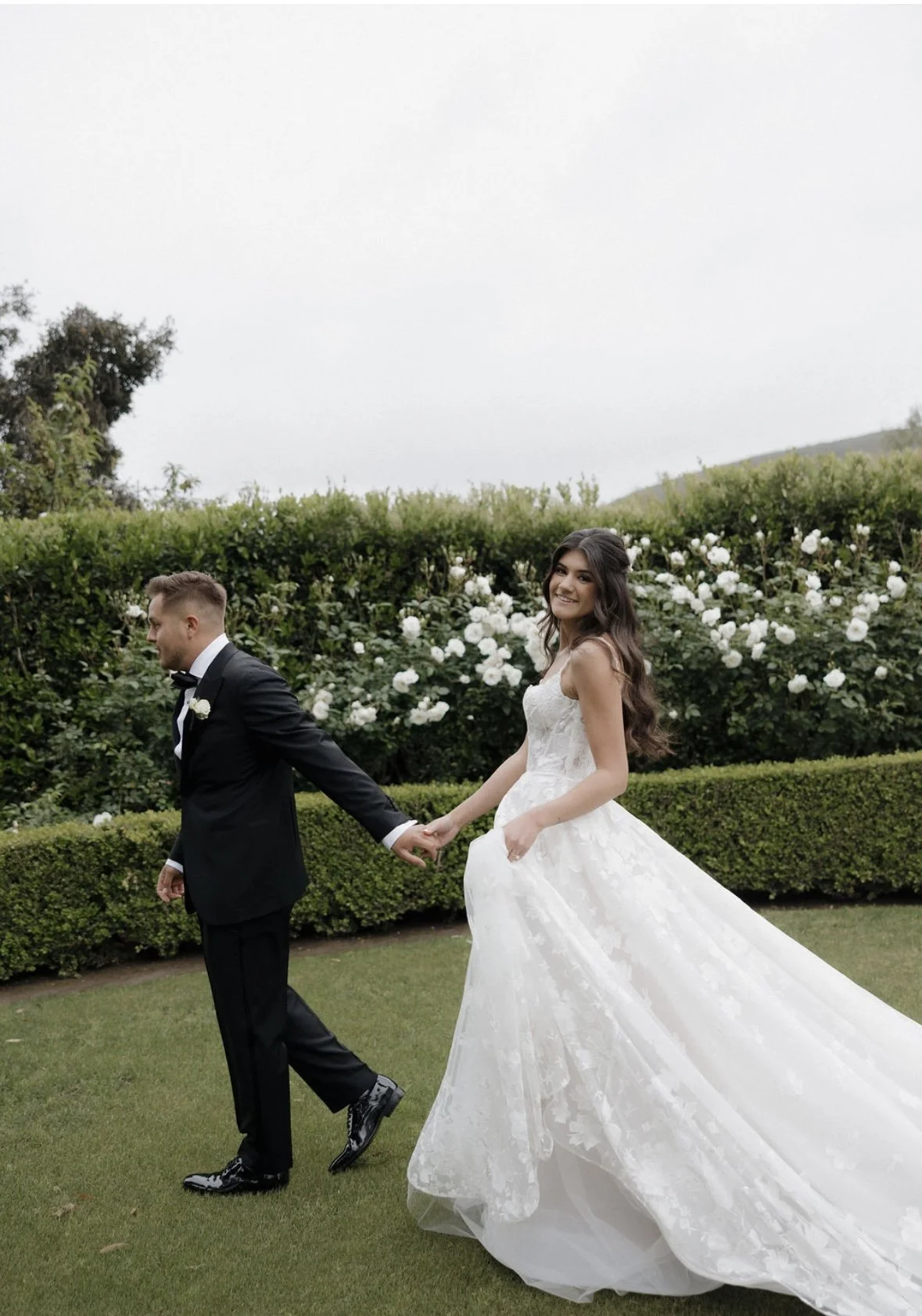 Bride and groom holding hands, walking on grass in a garden with white roses and green shrubs