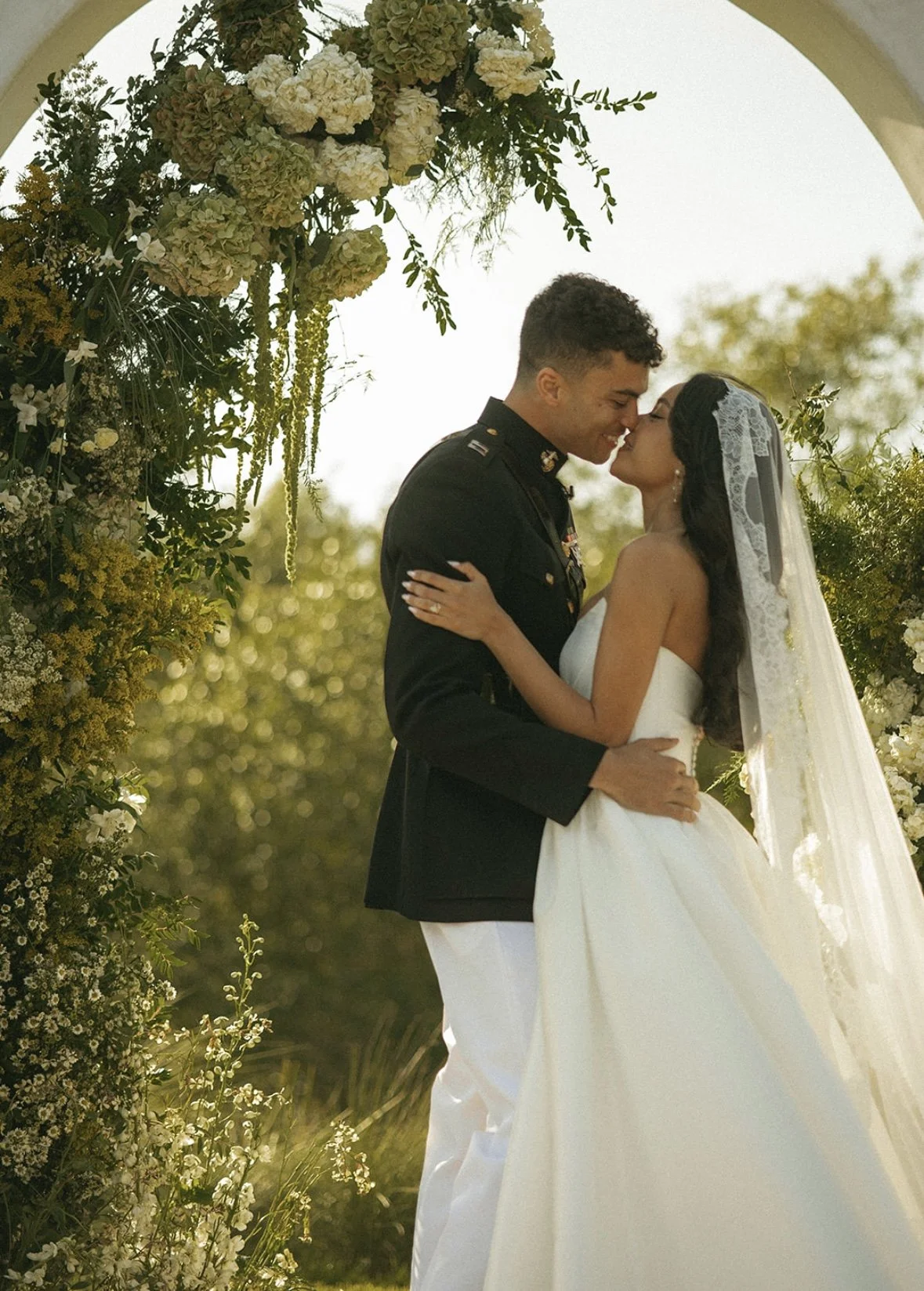 A bride and groom share a kiss during their outdoor wedding under a floral archway with white and green flowers, with sunlit trees in the background.