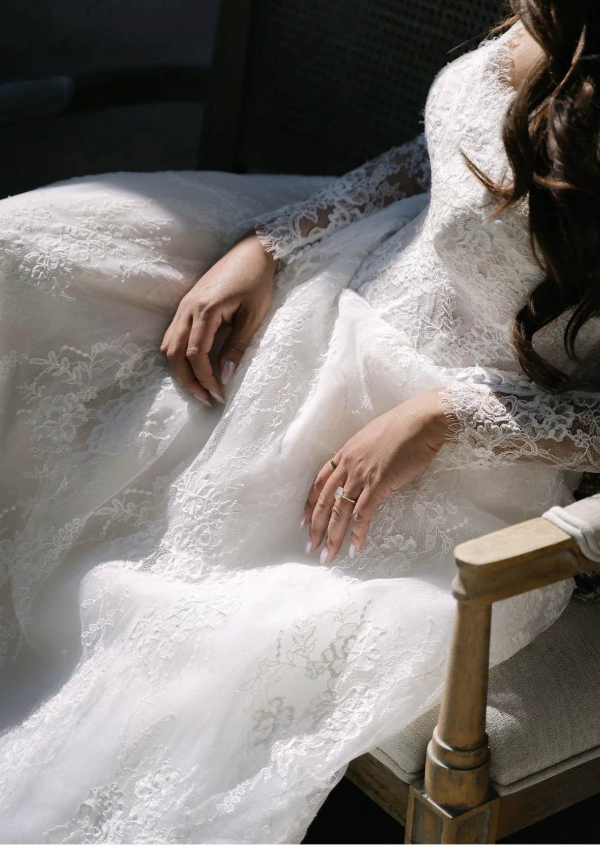 A bride in a white lace wedding dress sitting on a wooden bench, with her hands resting on her lap, showing a wedding ring.