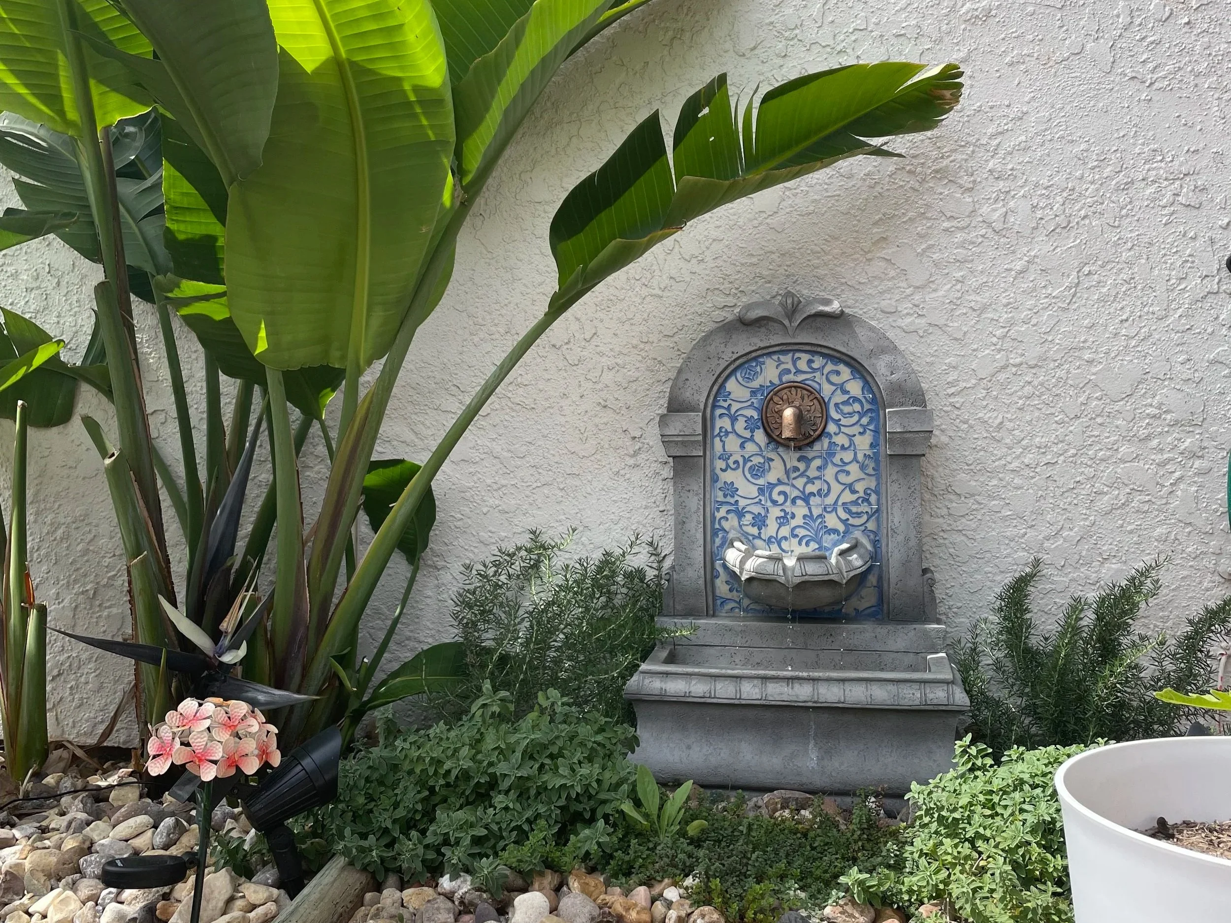 Small floor water fountain set against an exterior house wall surrounded by herbs