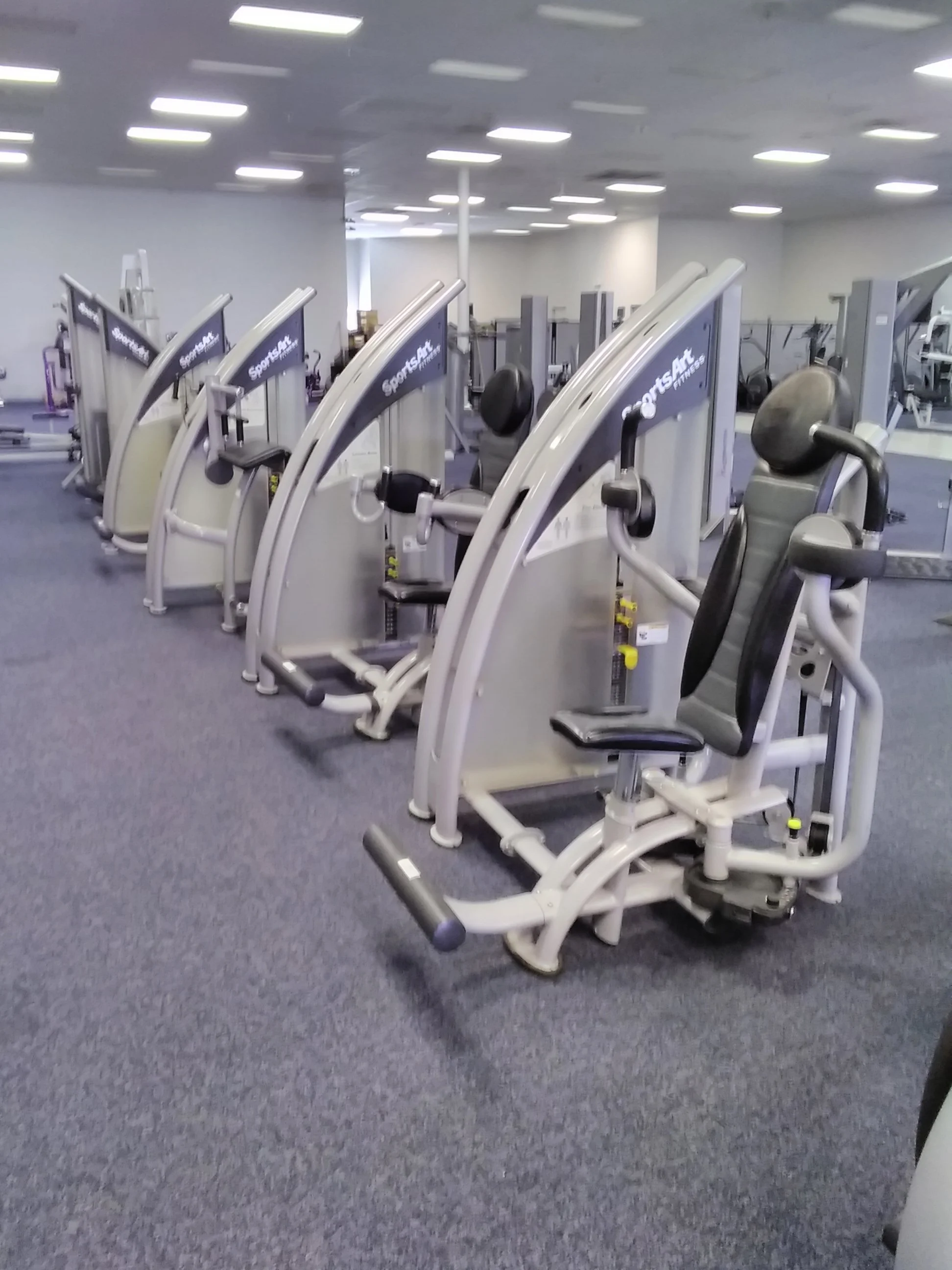 Row of exercise machines in a gym, each with black seats and handles, on a dark carpeted floor.