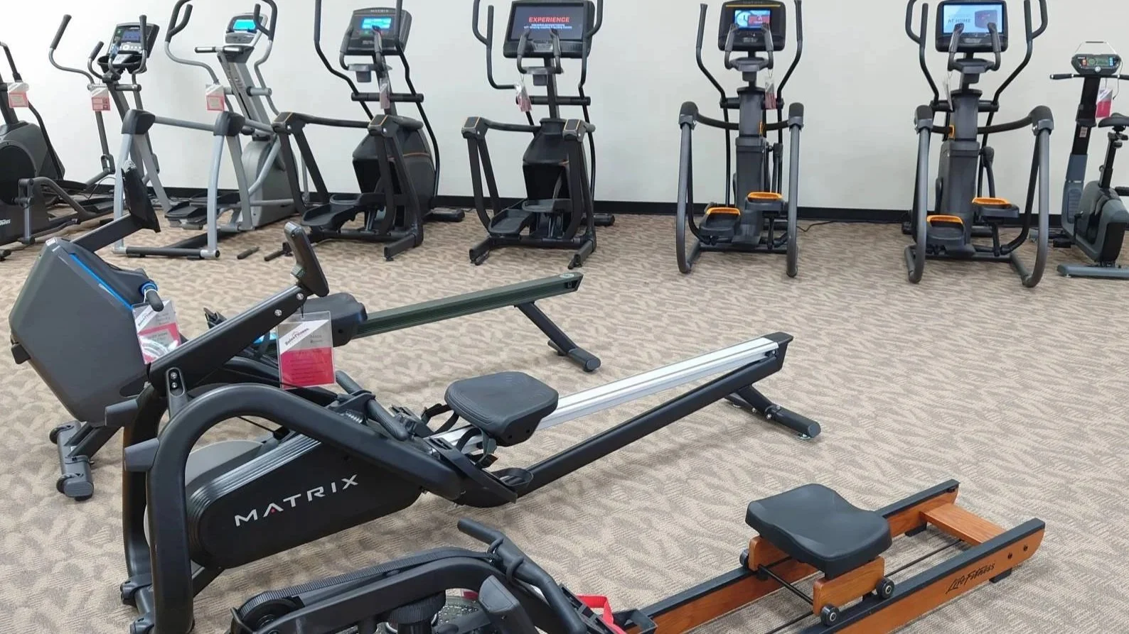 Row of various fitness equipment including elliptical machines, a rowing machine, and a stepper in a gym with beige carpeted floor.