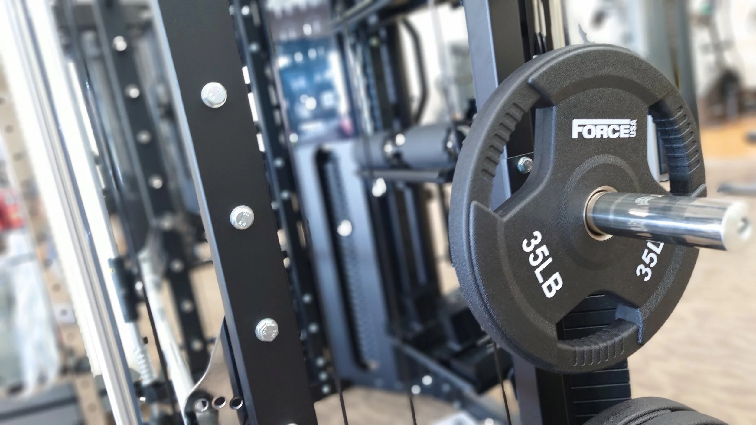 Close-up of a black 35-pound weight plate labeled 'FORCE USA 35 LB 35'. The weight plate is mounted on a metal barbell in a gym setting, with a metal weight rack in the background.