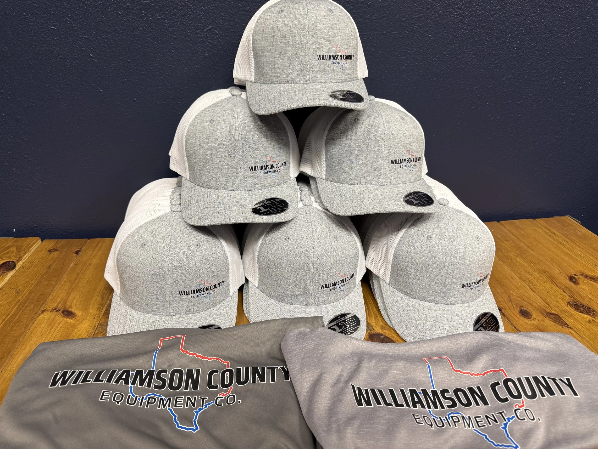 Stack of gray Williamson County Equipment Co. baseball caps with a logo featuring the outline of Texas in red and blue, overlapping stylized text, on a wooden surface against a dark wall.