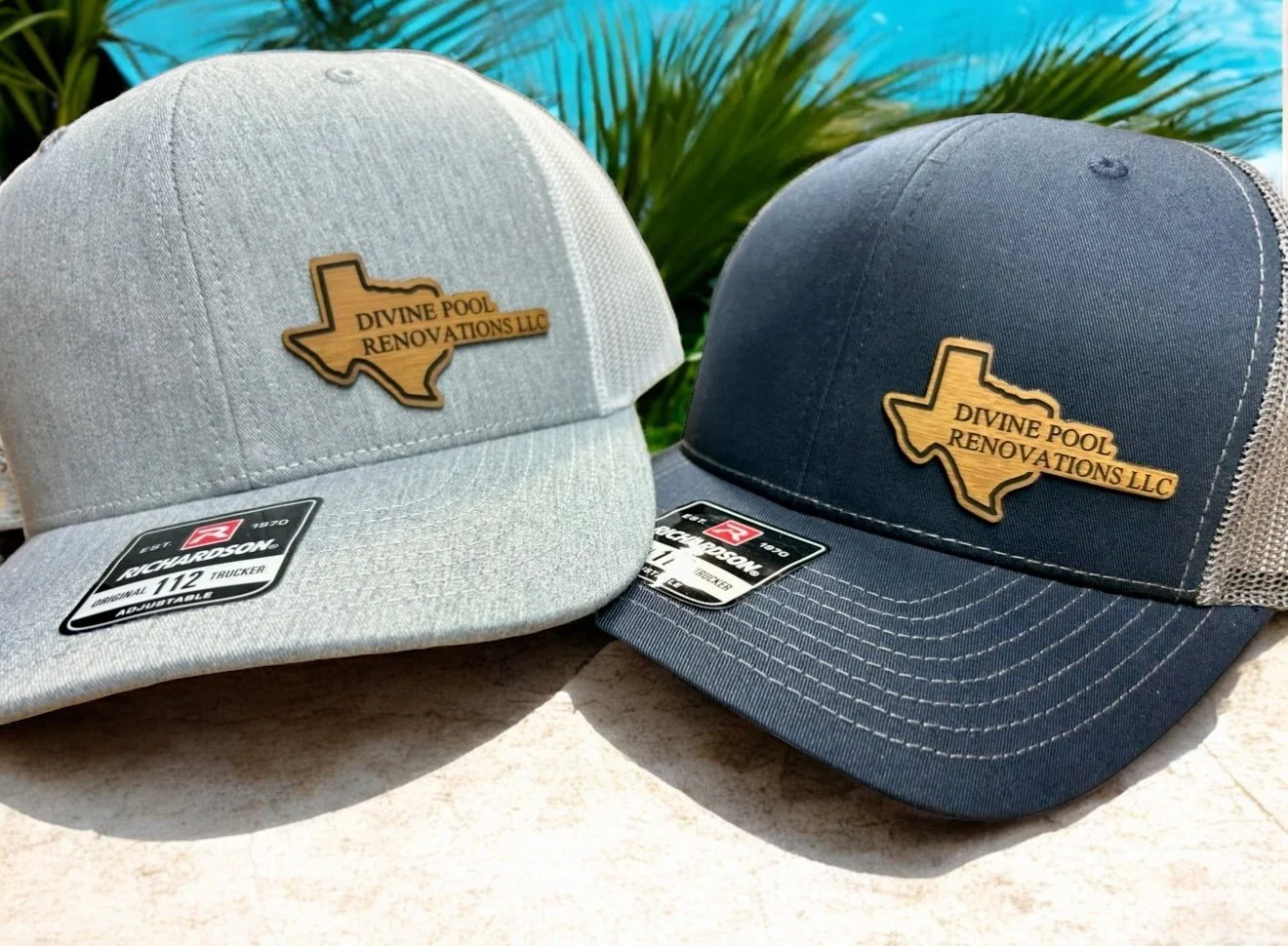 Two baseball caps with patches of Texas shape and the text "Divine Pool Renovations LLC" on a beige surface outdoors, with a background of green plants and a blue sky.