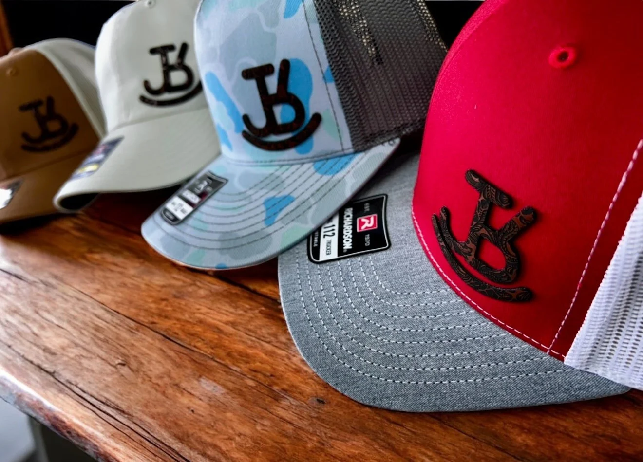 A collection of baseball caps with a logo featuring a smiling face and the letter J, arranged on a wooden surface. The caps have different colors and designs, including beige, blue camouflage, gray, and red, with a mesh back on one of the caps.
