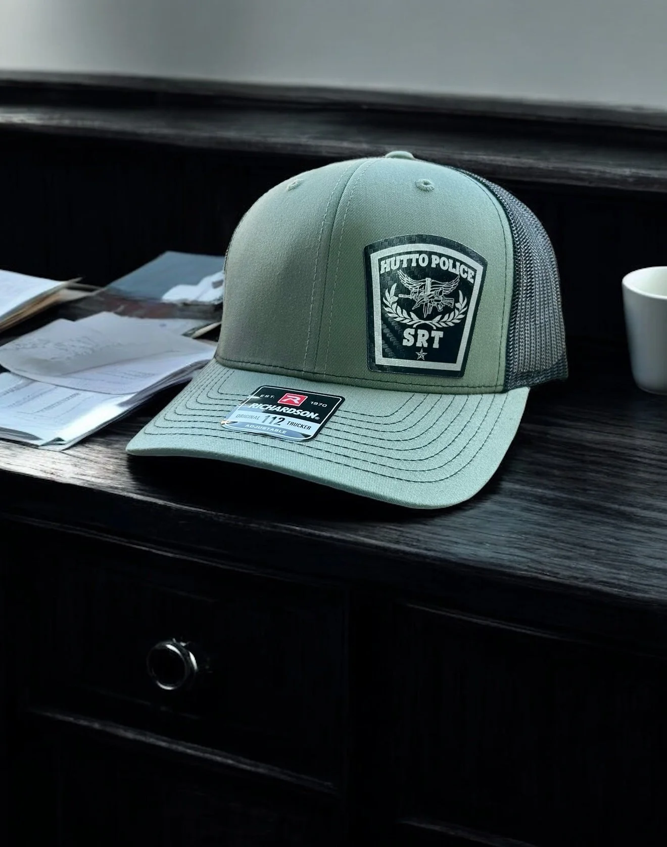 A green baseball cap with a patch that reads 'Hutto Police SRT' sitting on a dark wooden desk. There are papers, a white coffee mug, and a drawer with a round knob in the background.