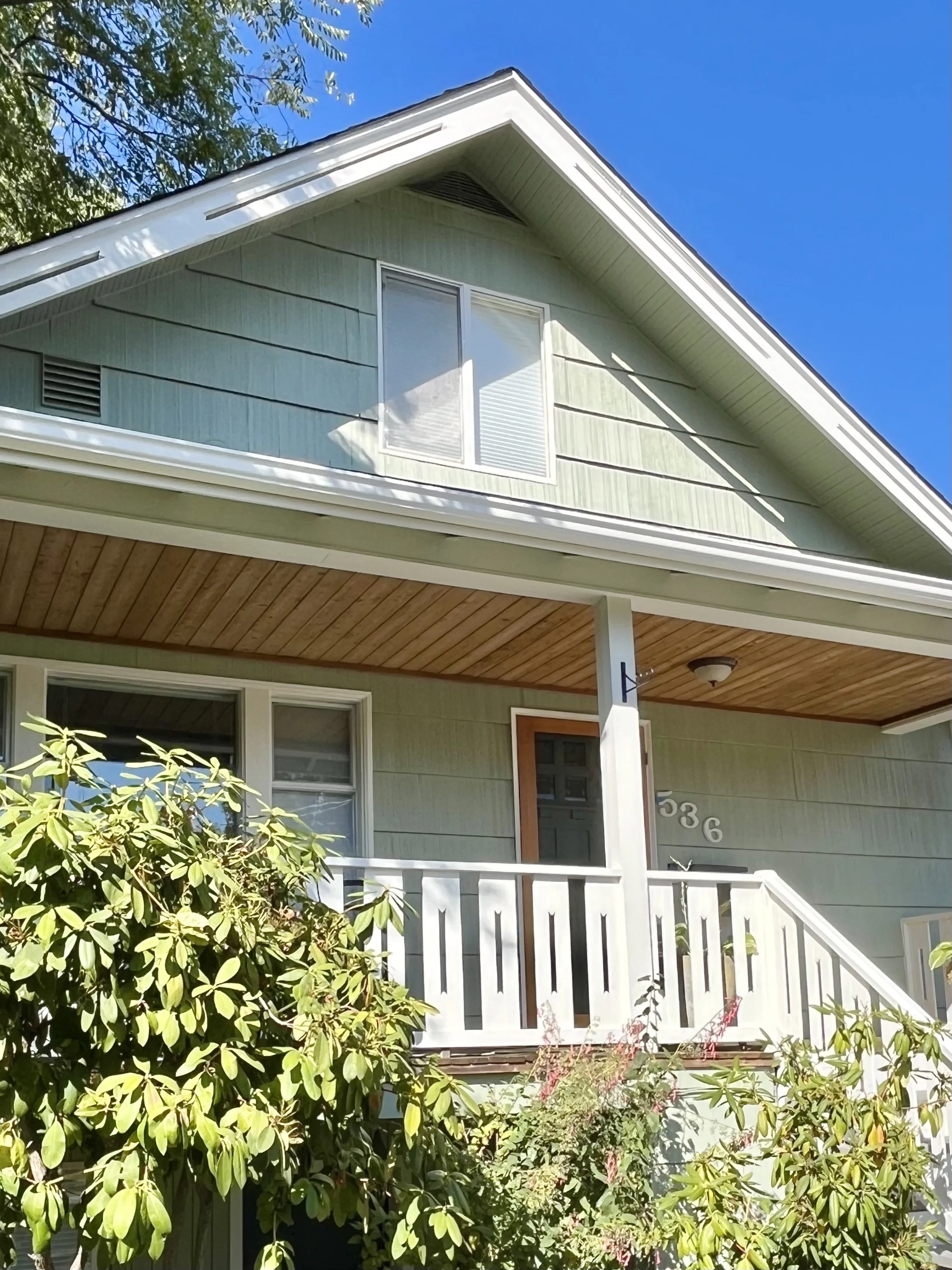 A sage green craftsman home with creamy white trim and a wood slat porch ceiling is angled in the sunlight.