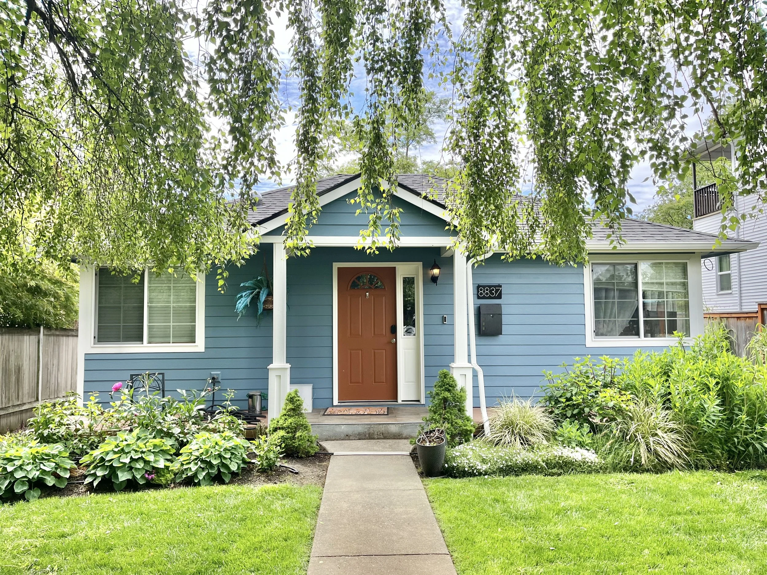 light blue house with white trim and a roasted brown front door color is shown with a beautiful garden in front.