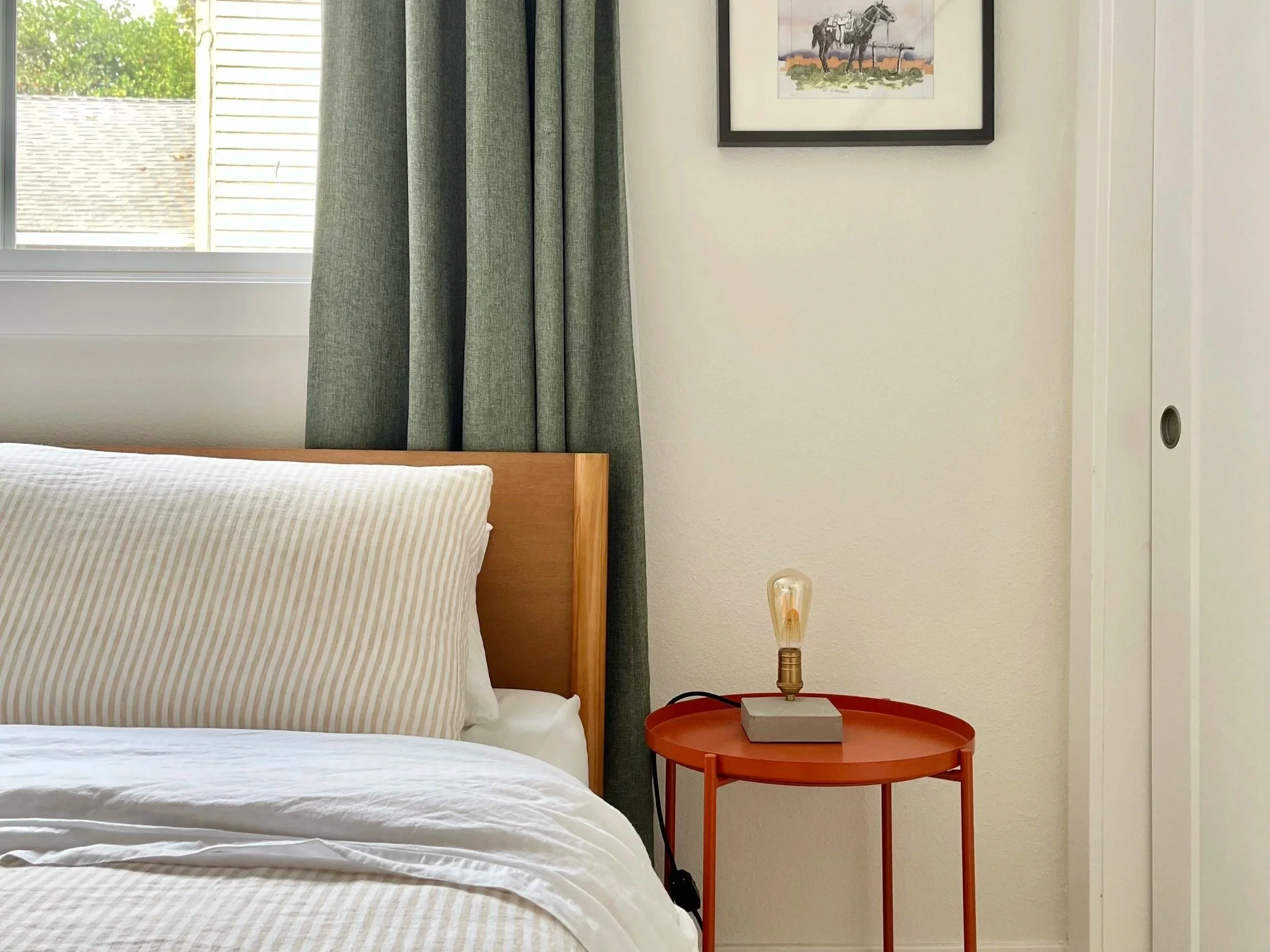 creamy off-white guest bedroom with soft tan and white striped bedding, sage green draperies, and a playful orange nightstand with a lightbulb lamp on top.