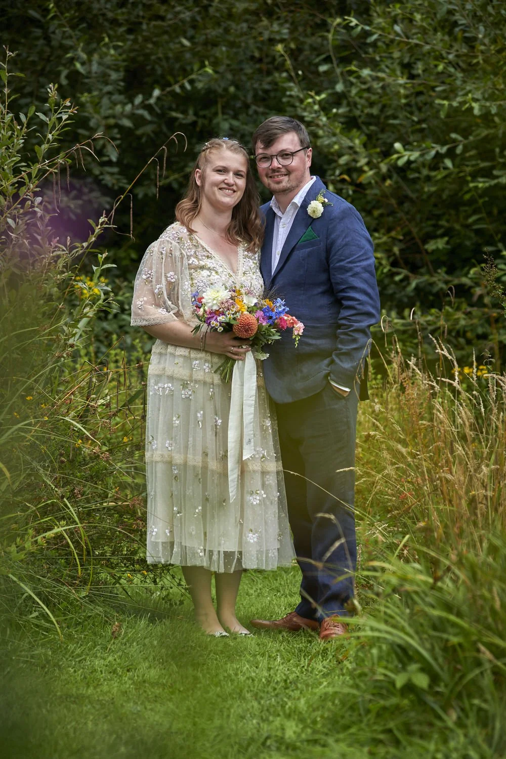 Bride and groom pose for full-length portrait, out-of-focus flowers are blurry in the foreground.